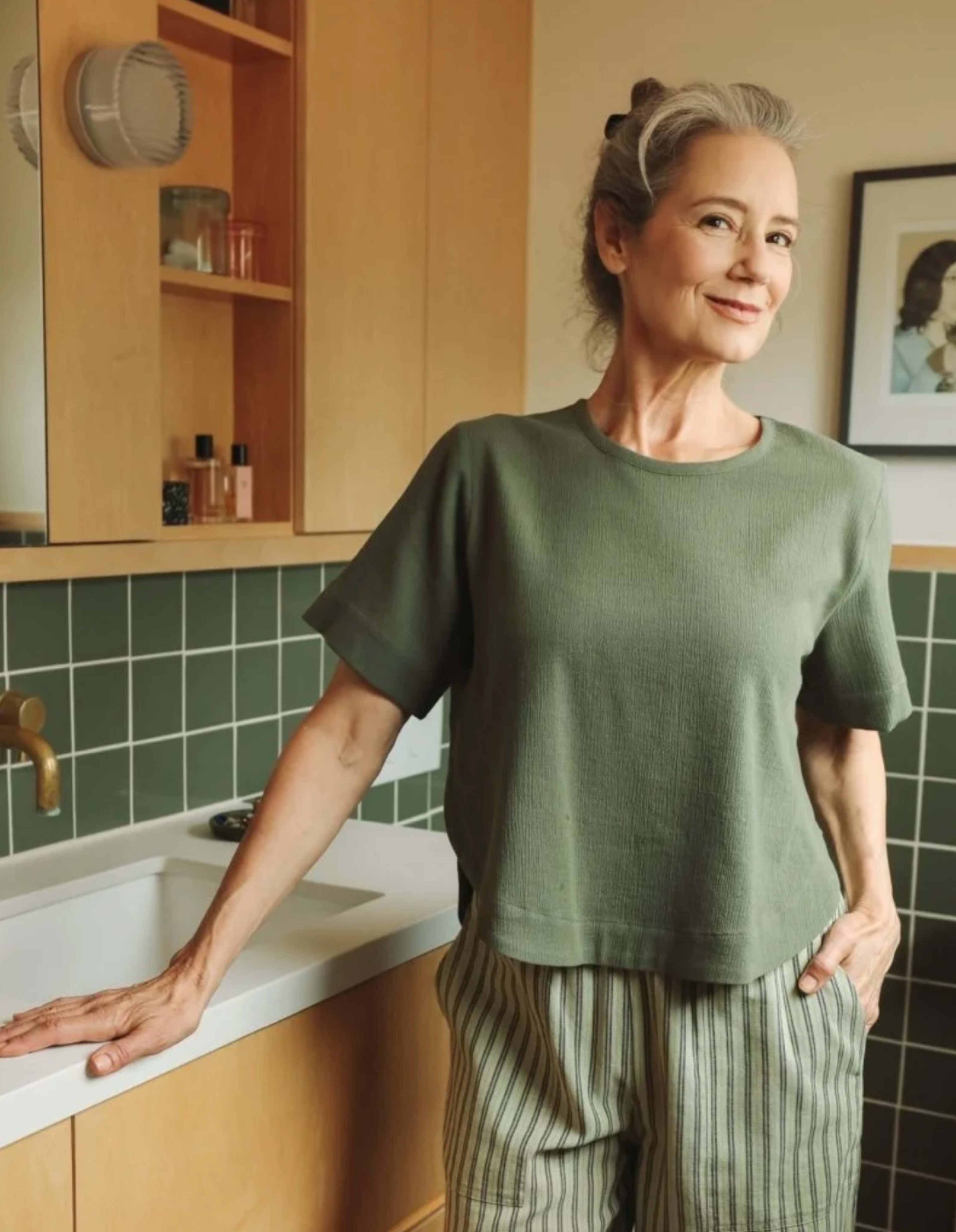 A woman in a green shirt and striped pants stands beside a sink in a modern bathroom, smiling at the camera.