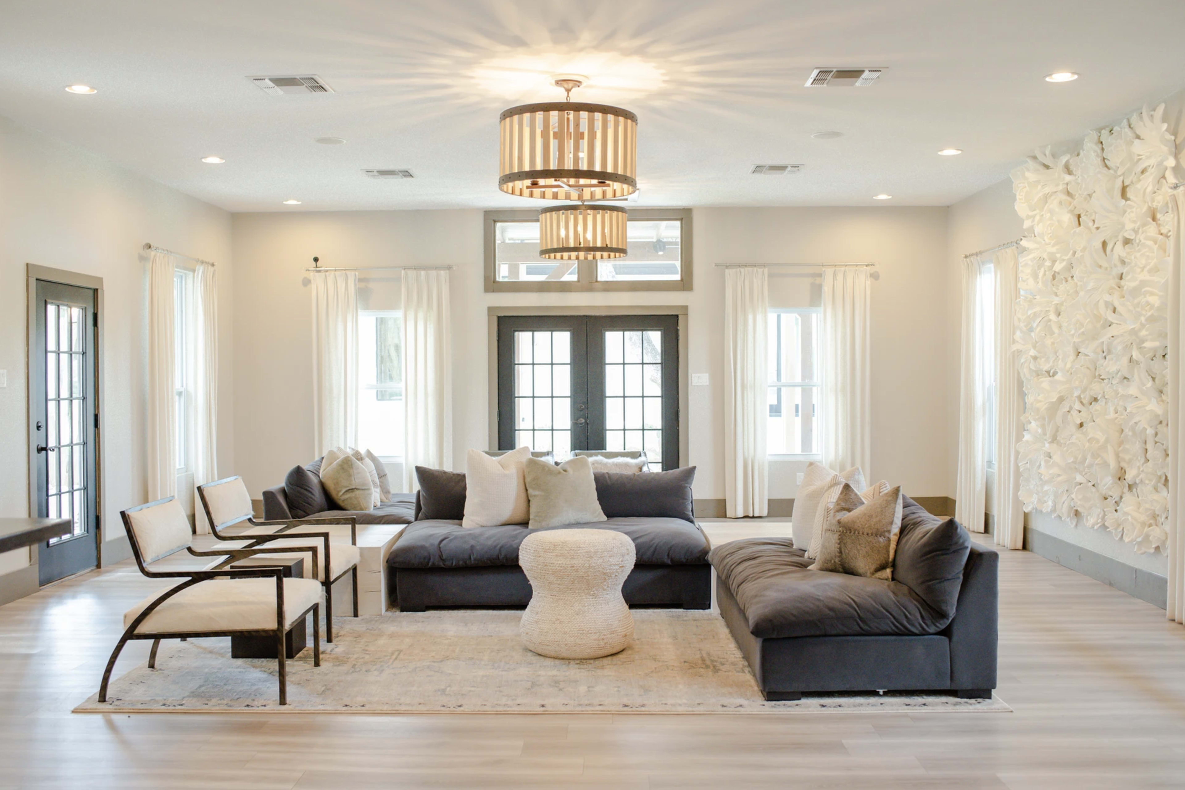 A modern living room features a dark sectional sofa, a round coffee table, and two chairs, surrounded by large windows and decorative wall accents.