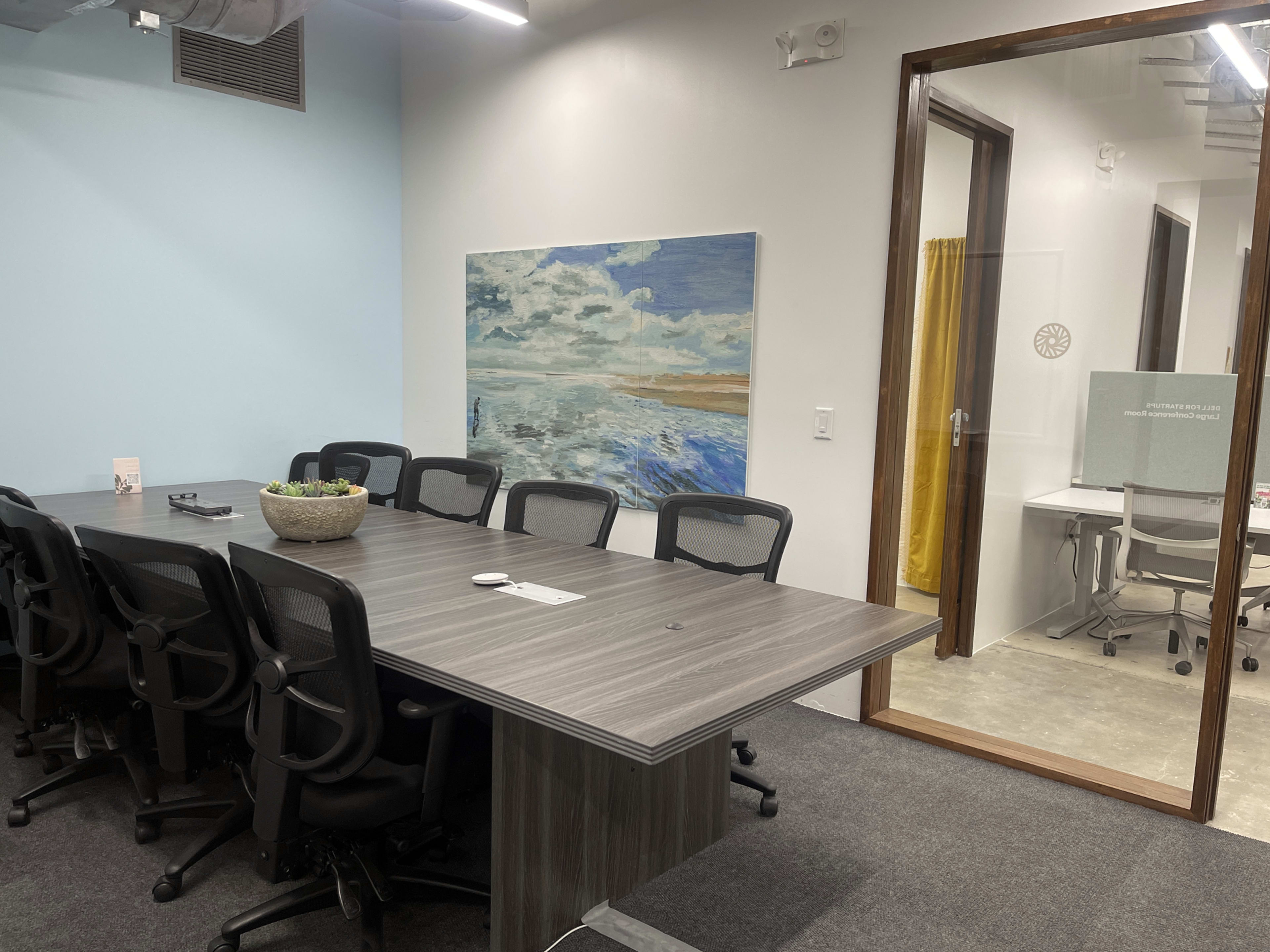 A conference room features a large gray table surrounded by ergonomic chairs, with a landscape painting on the wall and a glass door leading to a smaller meeting room.