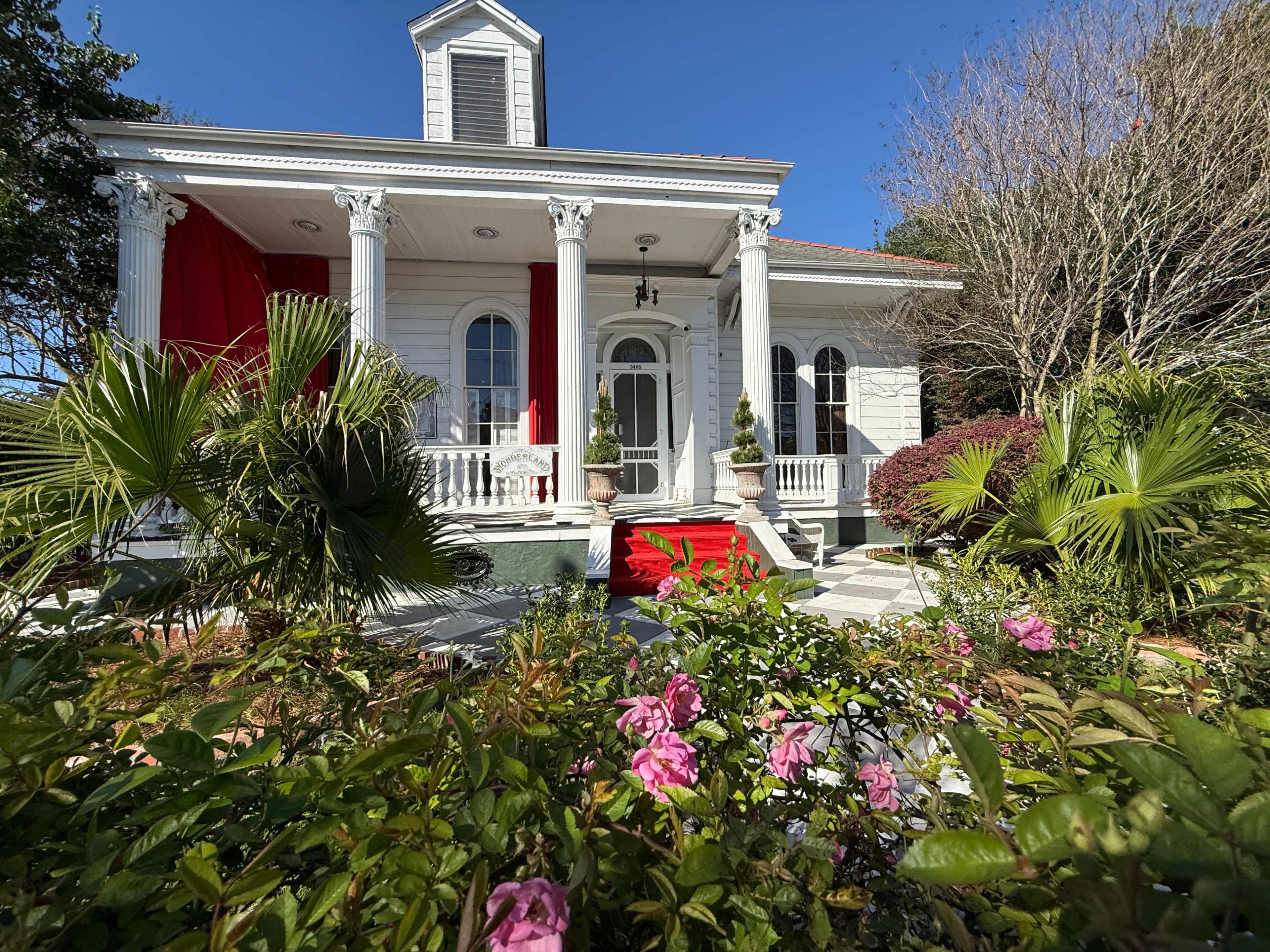 A stately white house with red accents features columns, a front porch, and surrounded by colorful flowers and greenery.