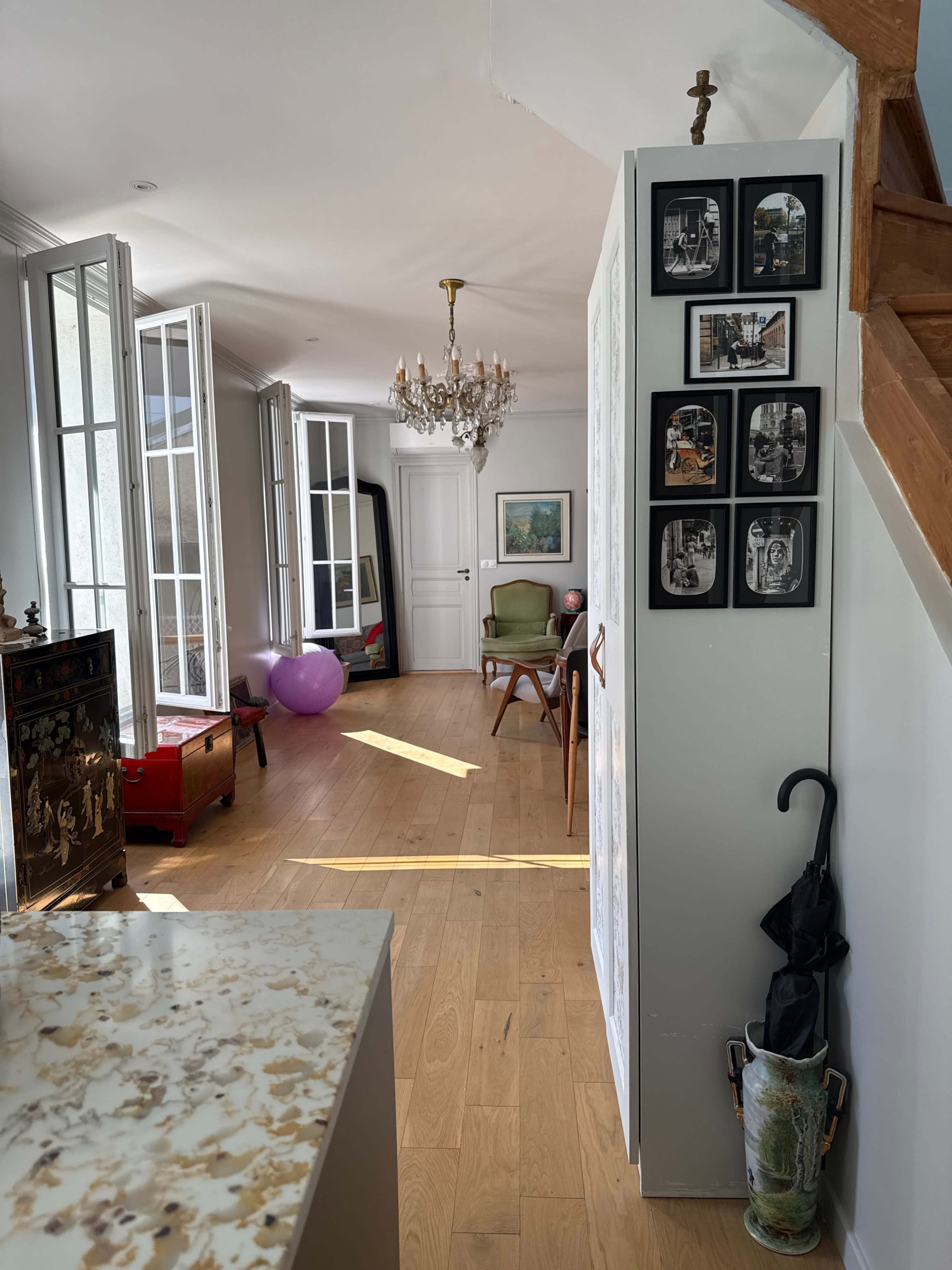 The image shows a bright, airy hallway with wooden flooring, a staircase, a chandelier, framed pictures on the wall, and furniture, including a chair and a painting in the background.
