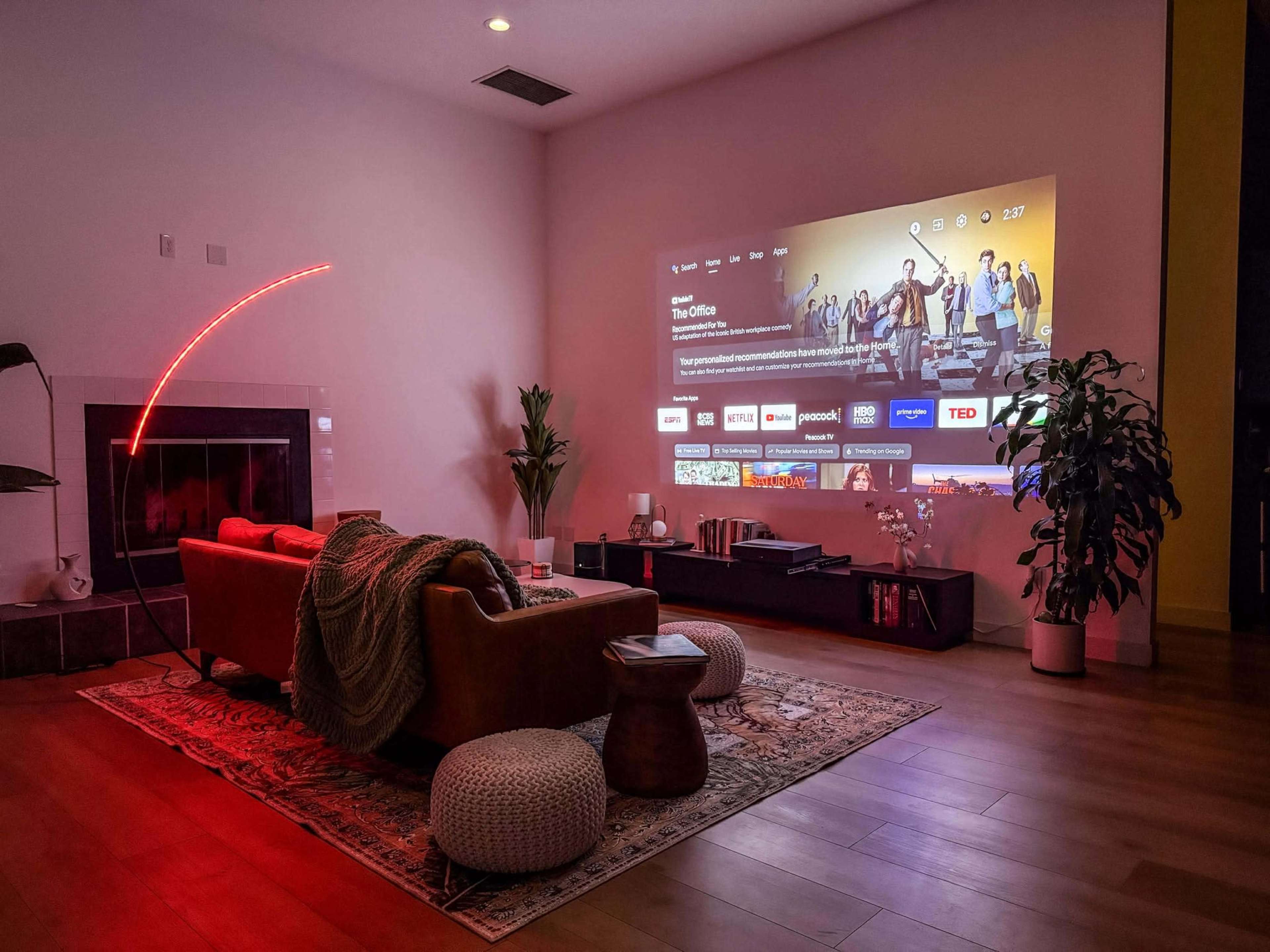A cozy living room is equipped with a large wall-mounted projector displaying a streaming service interface, featuring the show "The Office," alongside a comfortable couch and decorative plants.