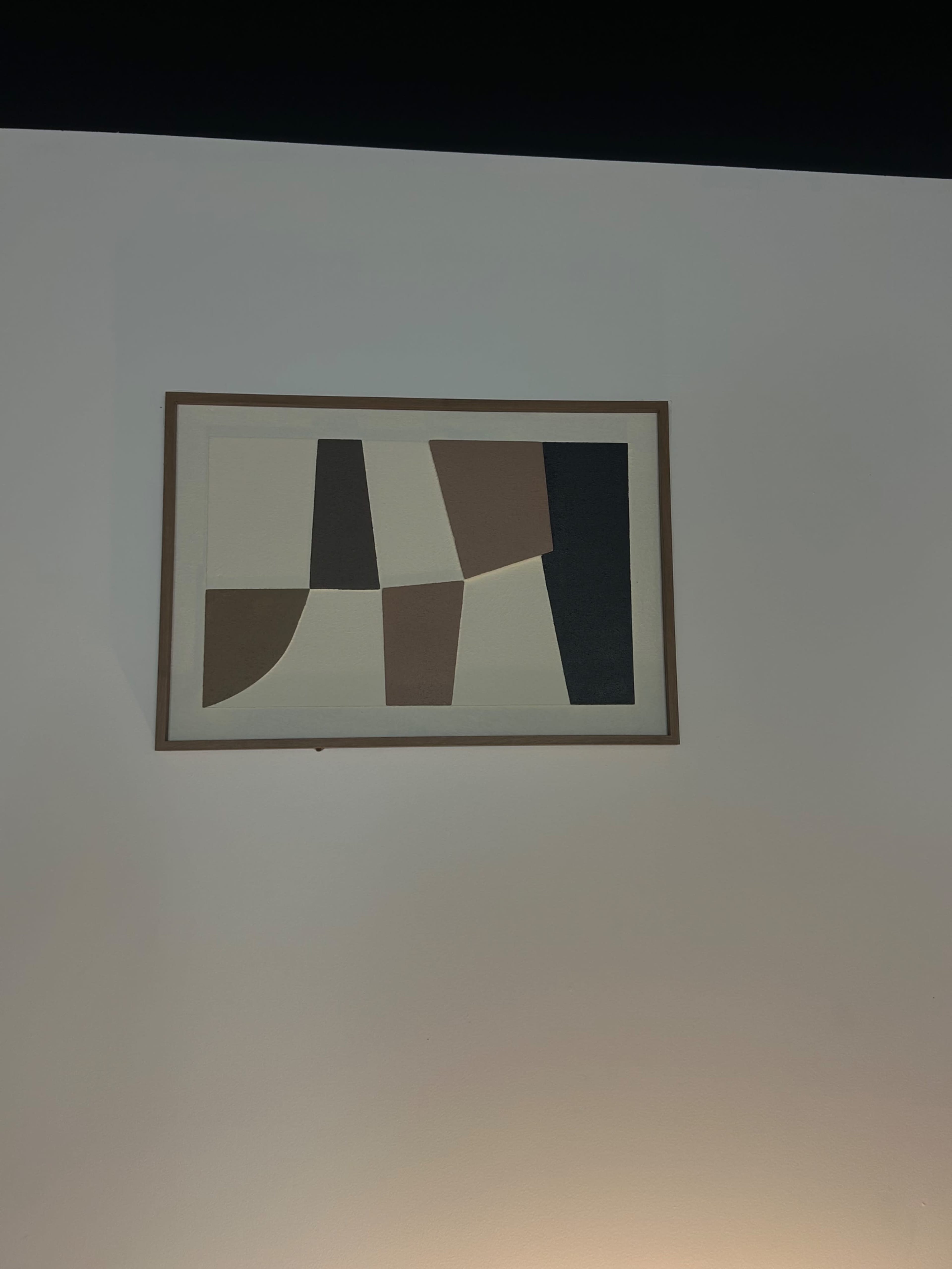 A framed abstract artwork featuring geometric shapes in various shades hangs on a white wall.