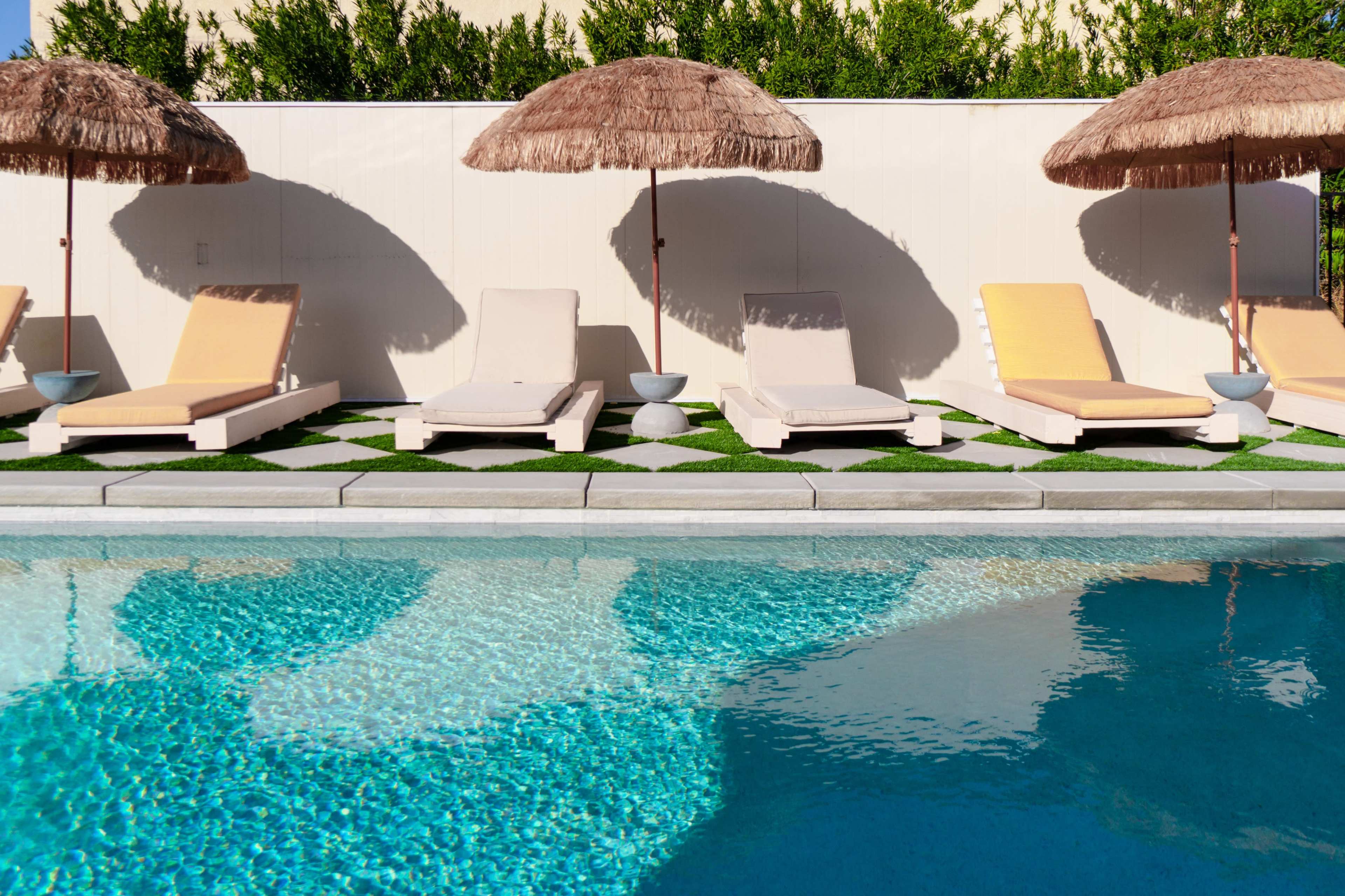 Lounge chairs with umbrellas are arranged beside a swimming pool surrounded by neatly maintained grass and a white wall.
