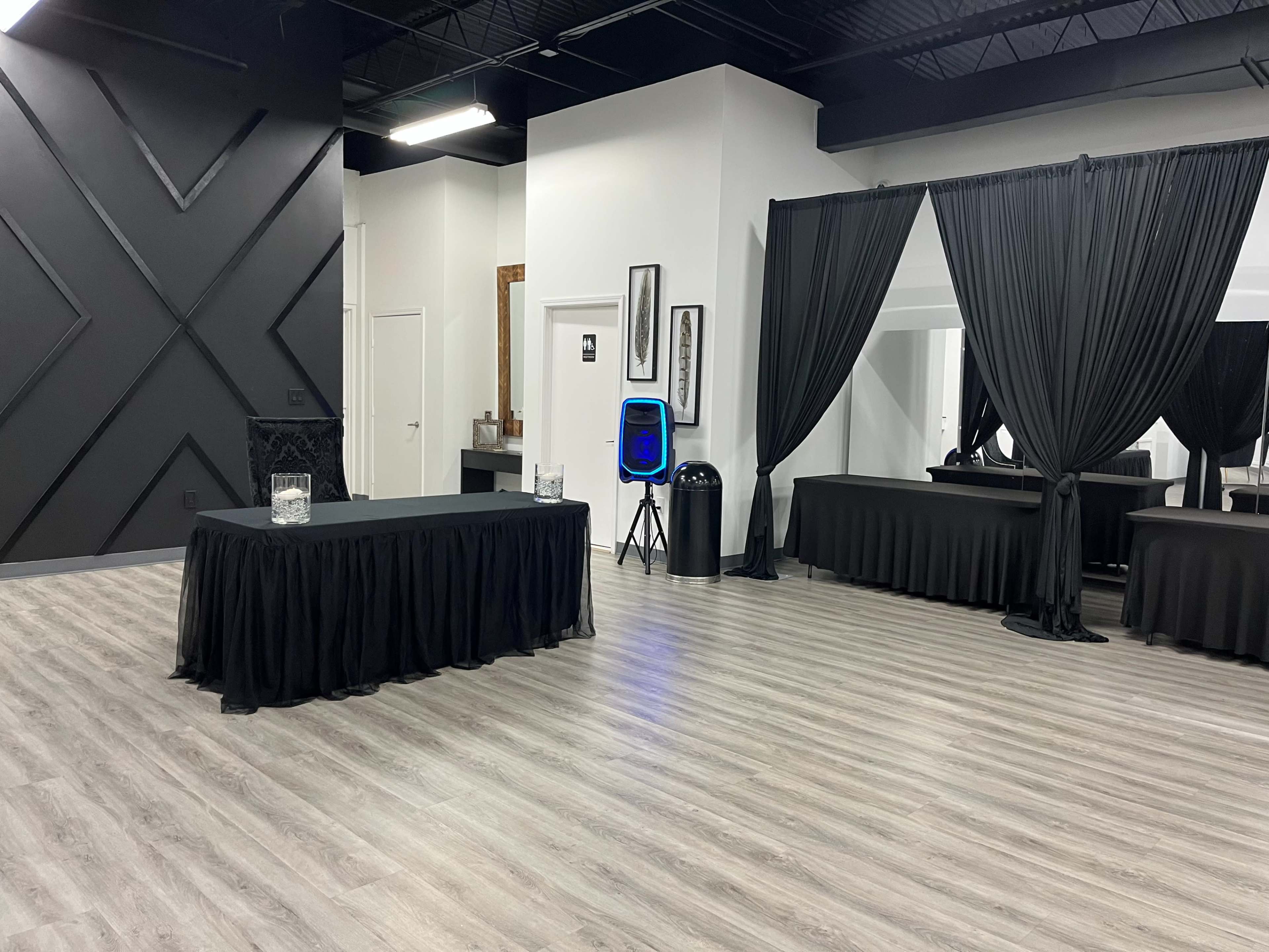 The image shows a modern interior space featuring several tables covered with black tablecloths and draped black curtains against a textured wall.