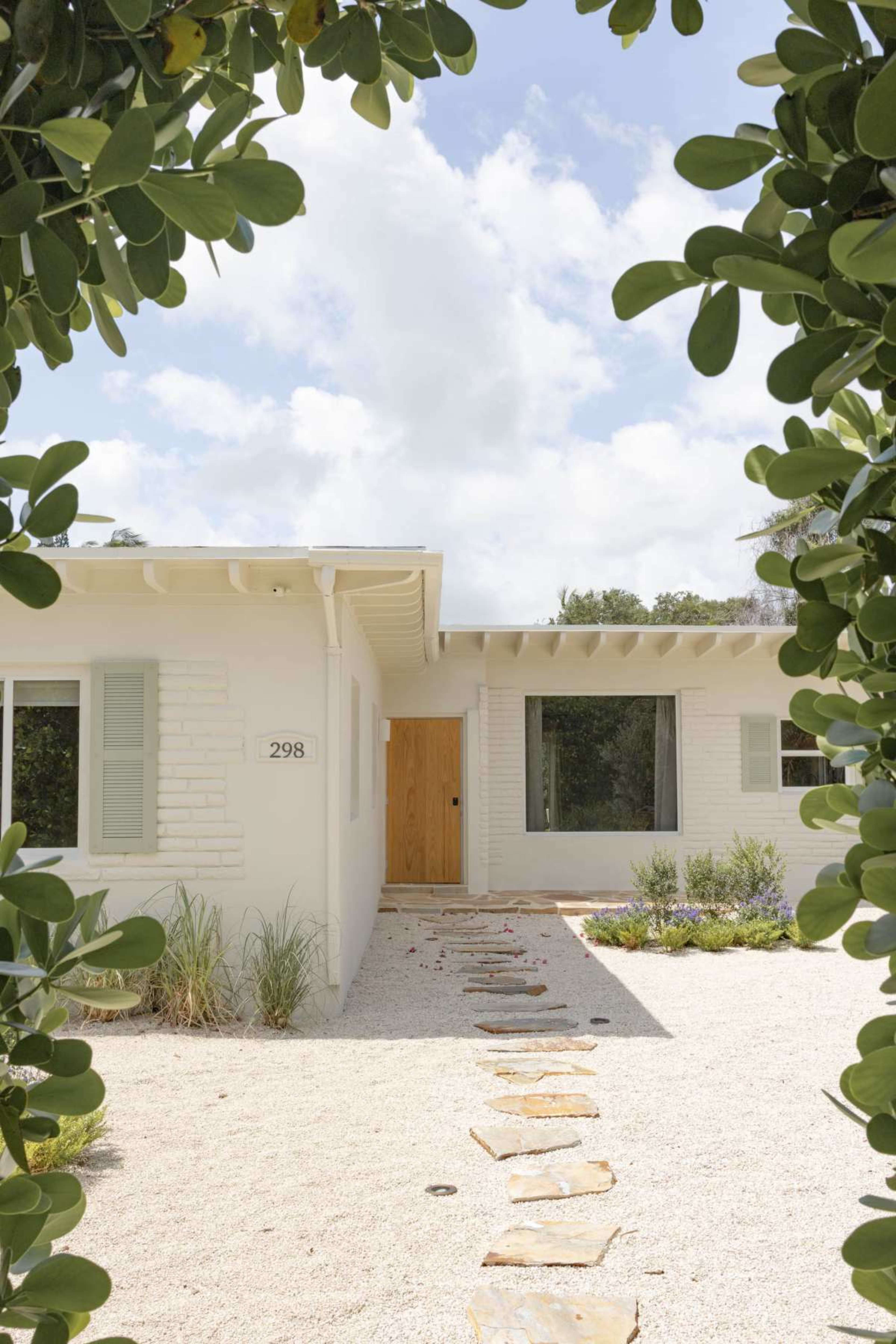 A white, single-story house with a wooden front door and a stone pathway is framed by green foliage.