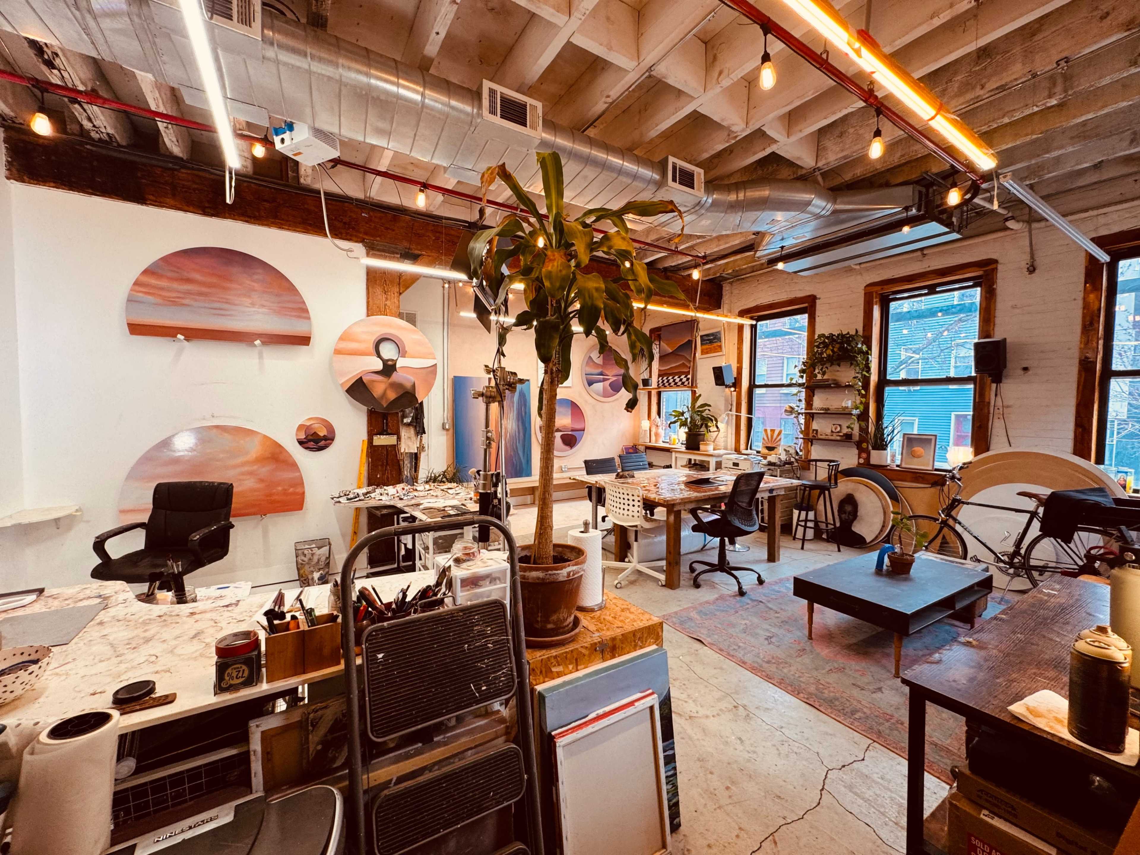 The image shows a spacious art studio featuring a desk, plants, various art supplies, and large round artworks on the walls.