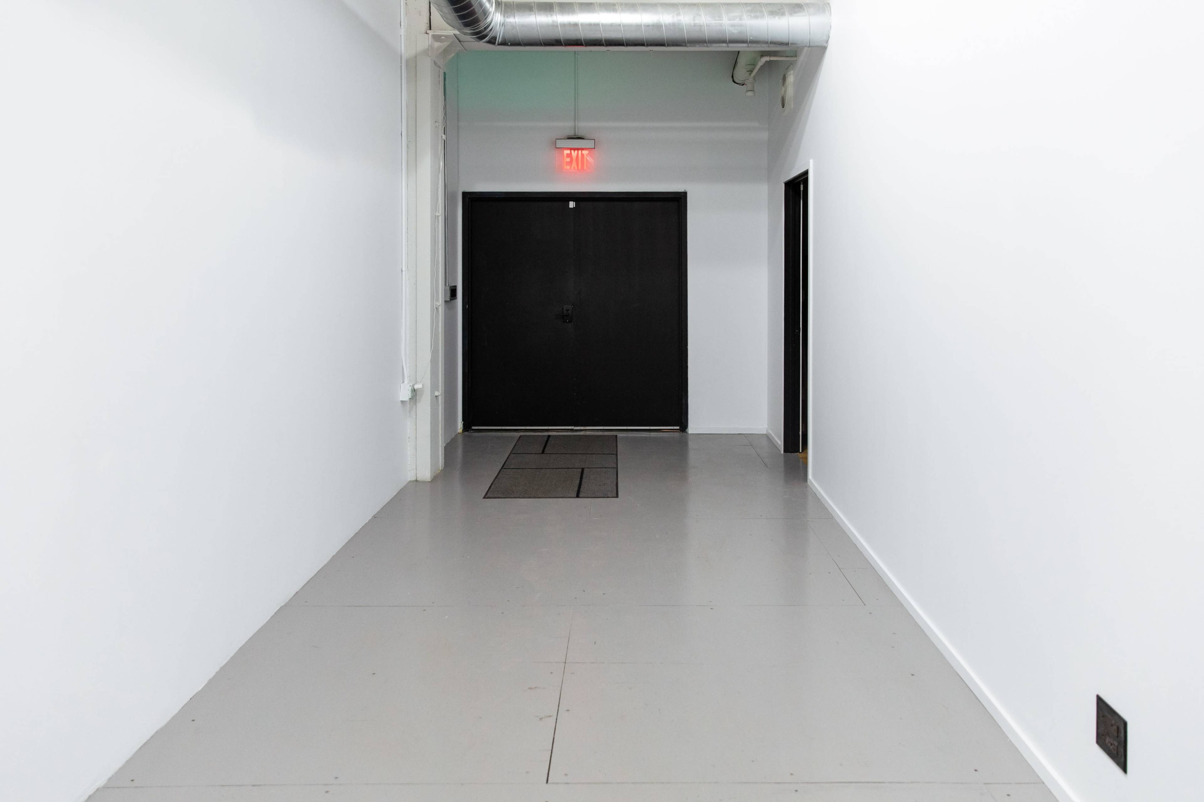 The image shows a narrow, well-lit hallway leading to a black door at the end, with a second door on the right side.