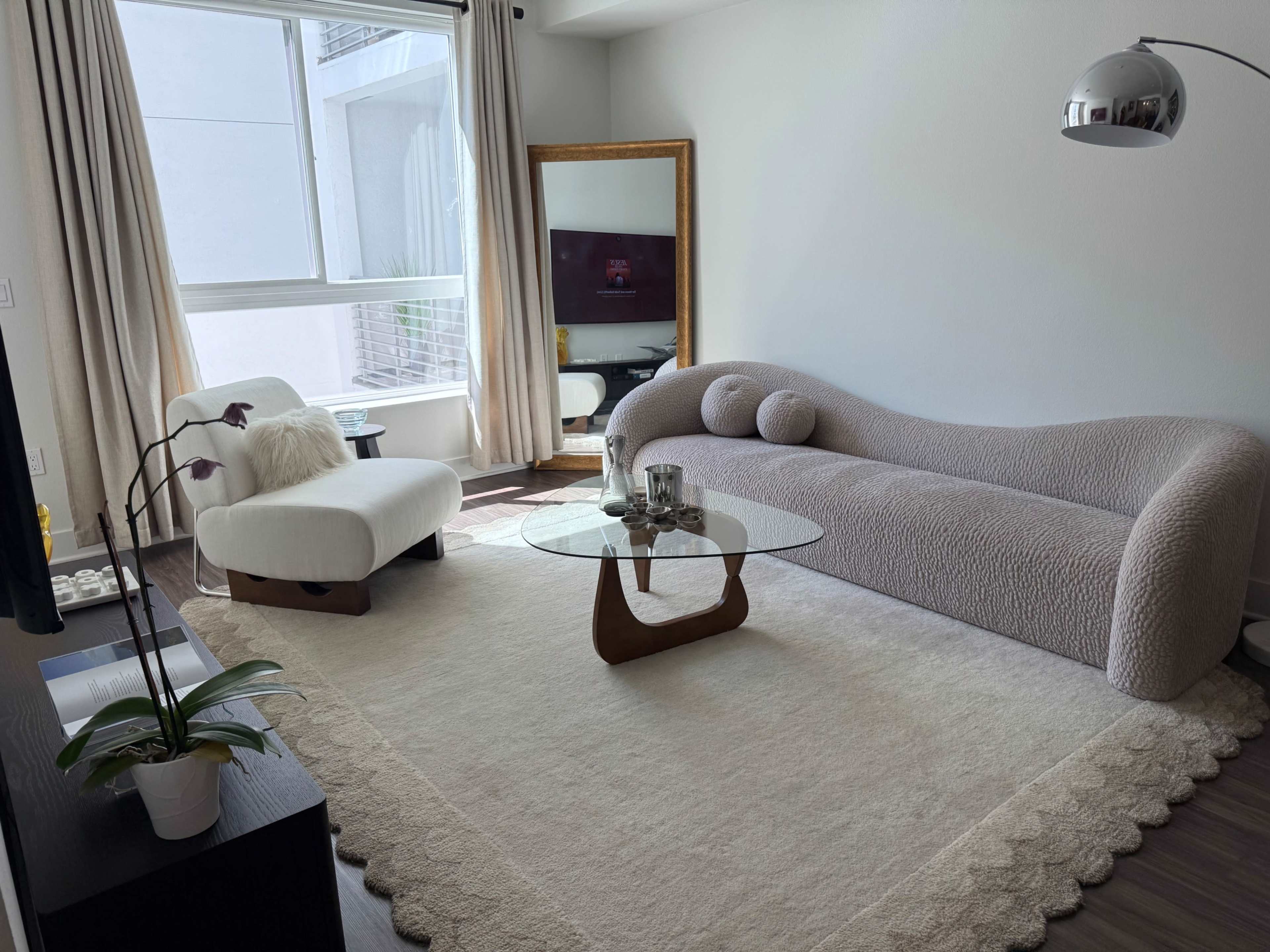 The image shows a modern living room featuring a curved beige sofa, a round glass coffee table, an armchair, a large mirror, and a television on a wooden stand.