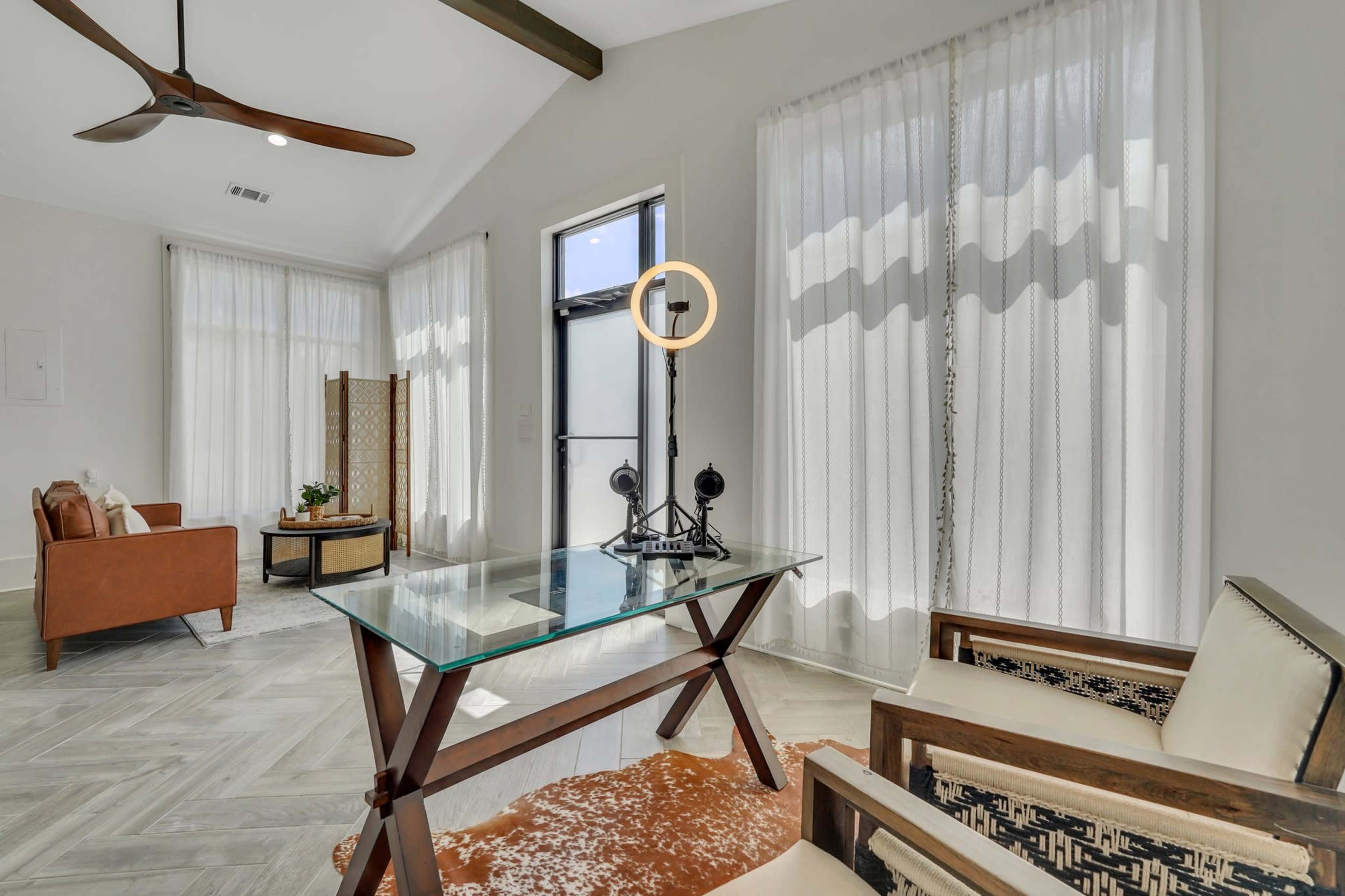 A modern living room features a glass table, wooden beams, and large windows with sheer curtains.