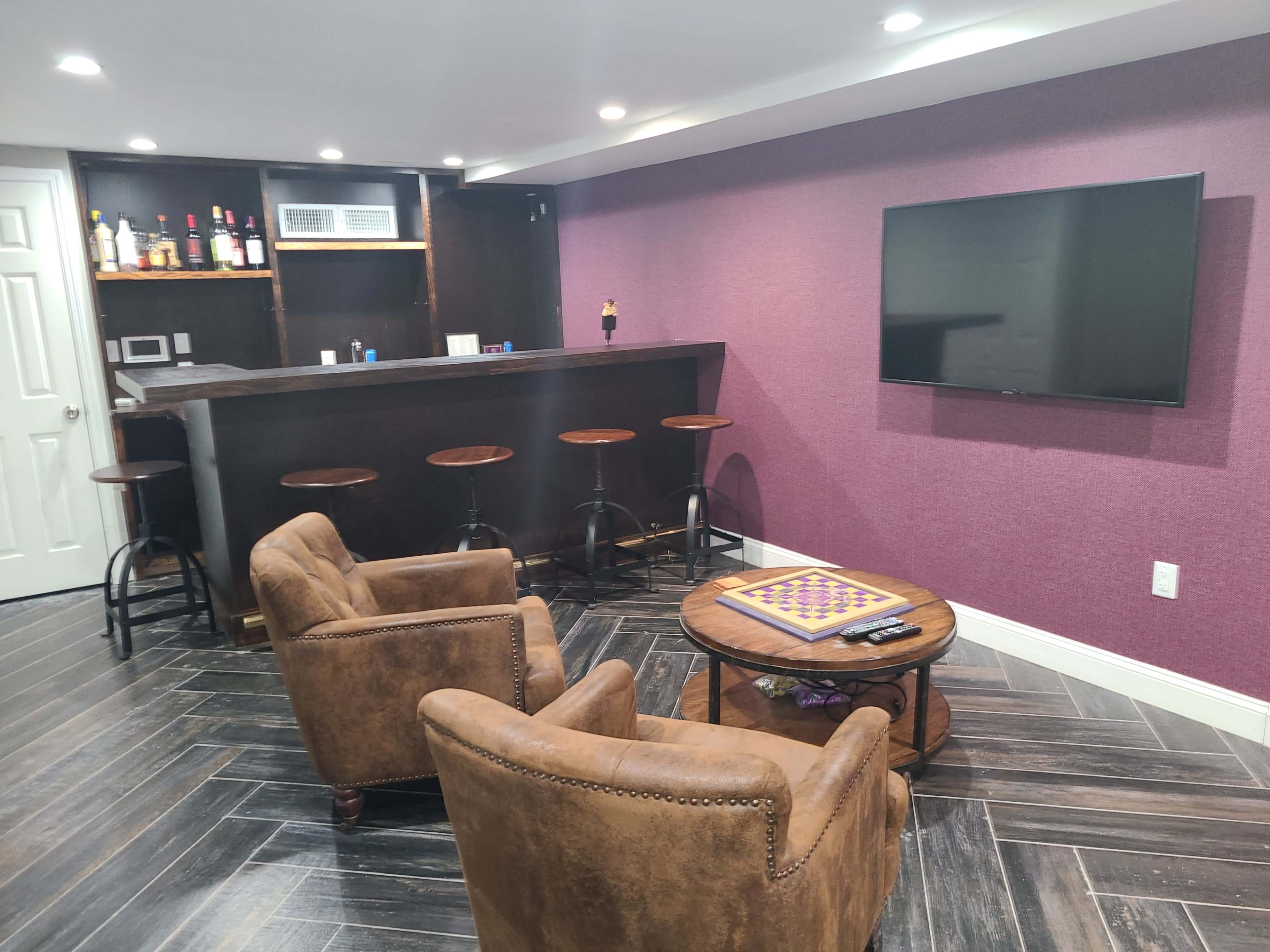 A cozy basement lounge features a bar with high stools, two brown armchairs, a round table with a game board, and a large flat-screen TV mounted on a purple wall.