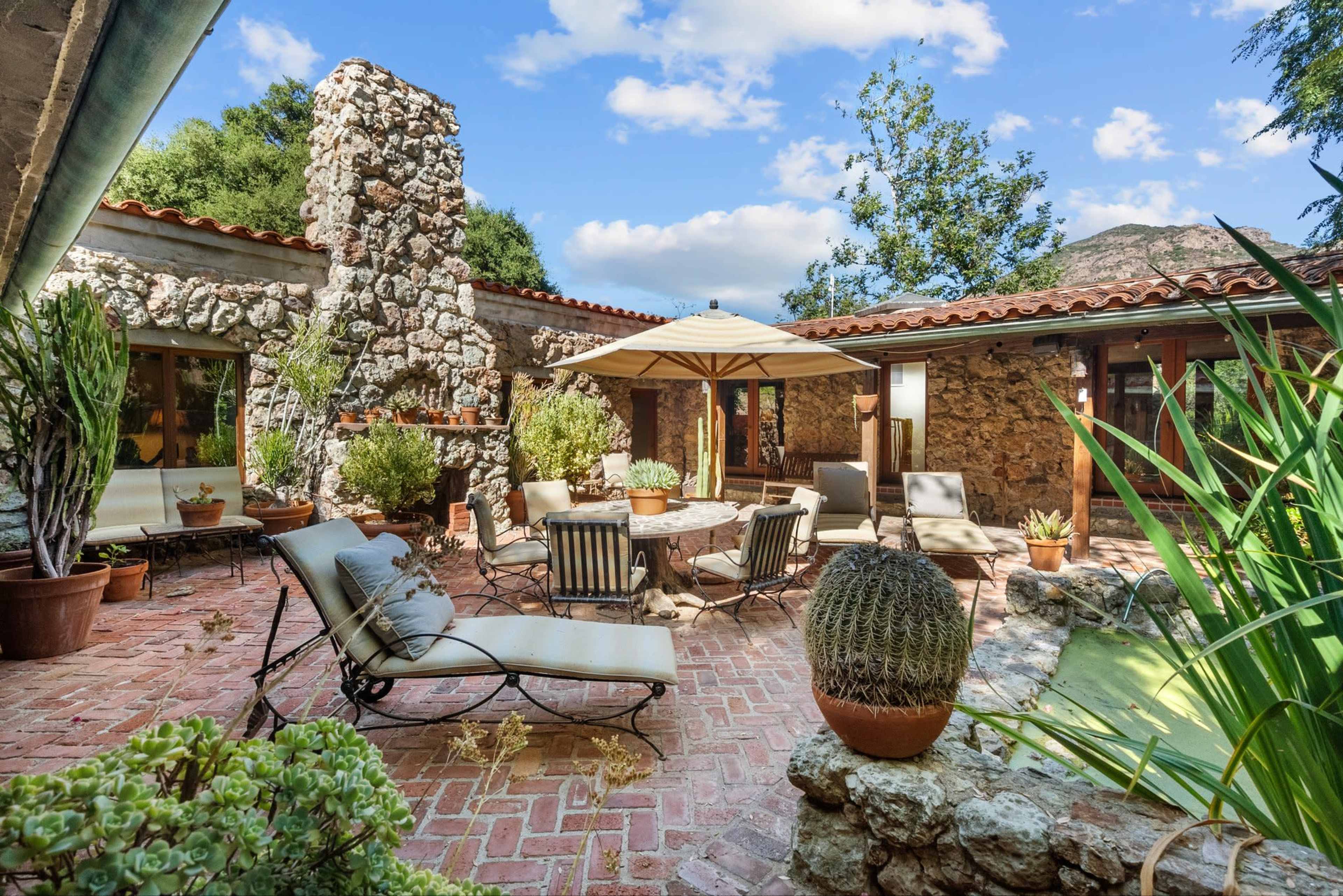 A stone courtyard features lounge chairs, an umbrella, and potted plants surrounded by a rustic stone building and greenery.