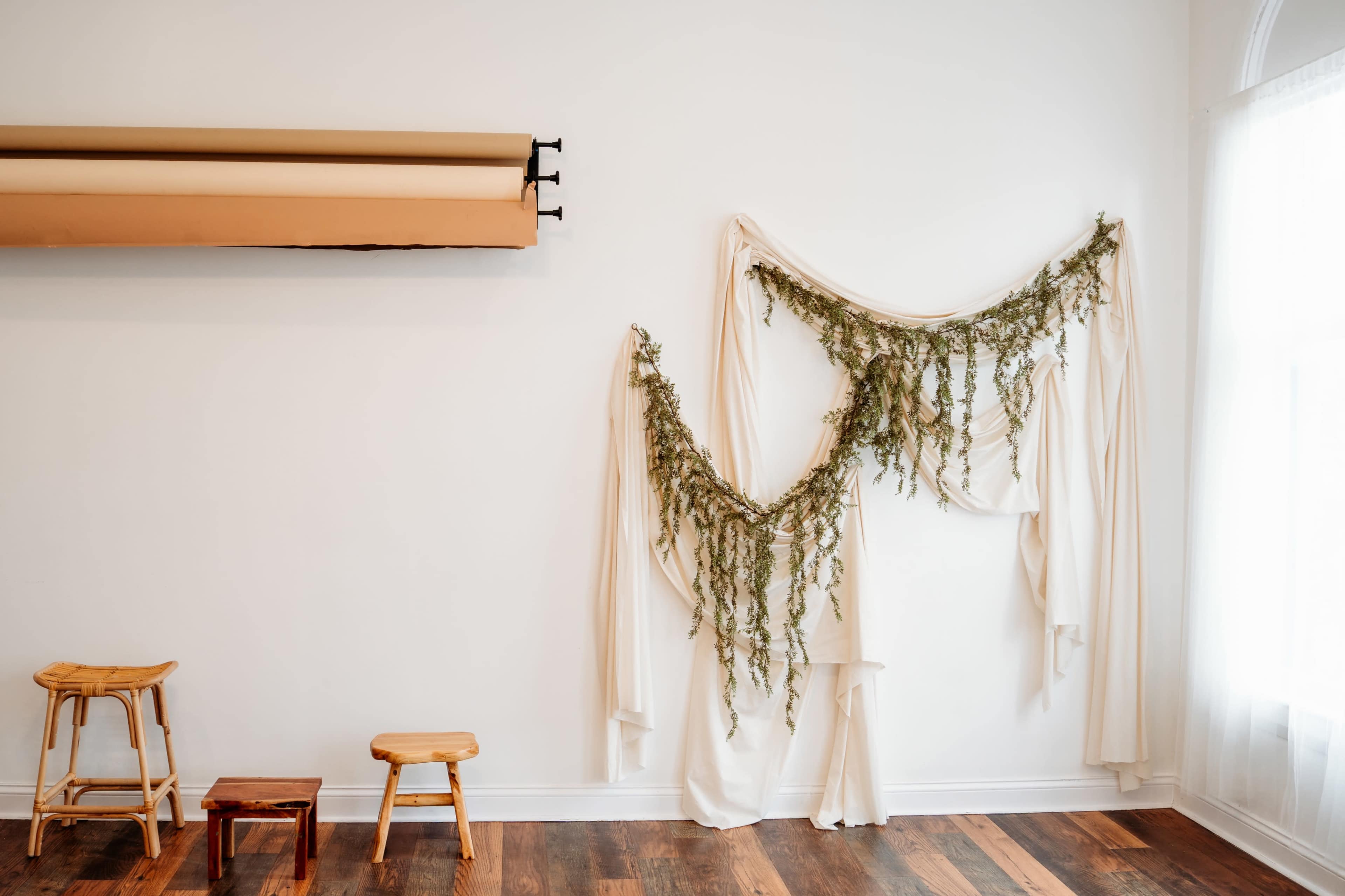 The image shows a minimalist interior featuring a white wall adorned with draped fabric and greenery, along with wooden stools positioned on the floor.