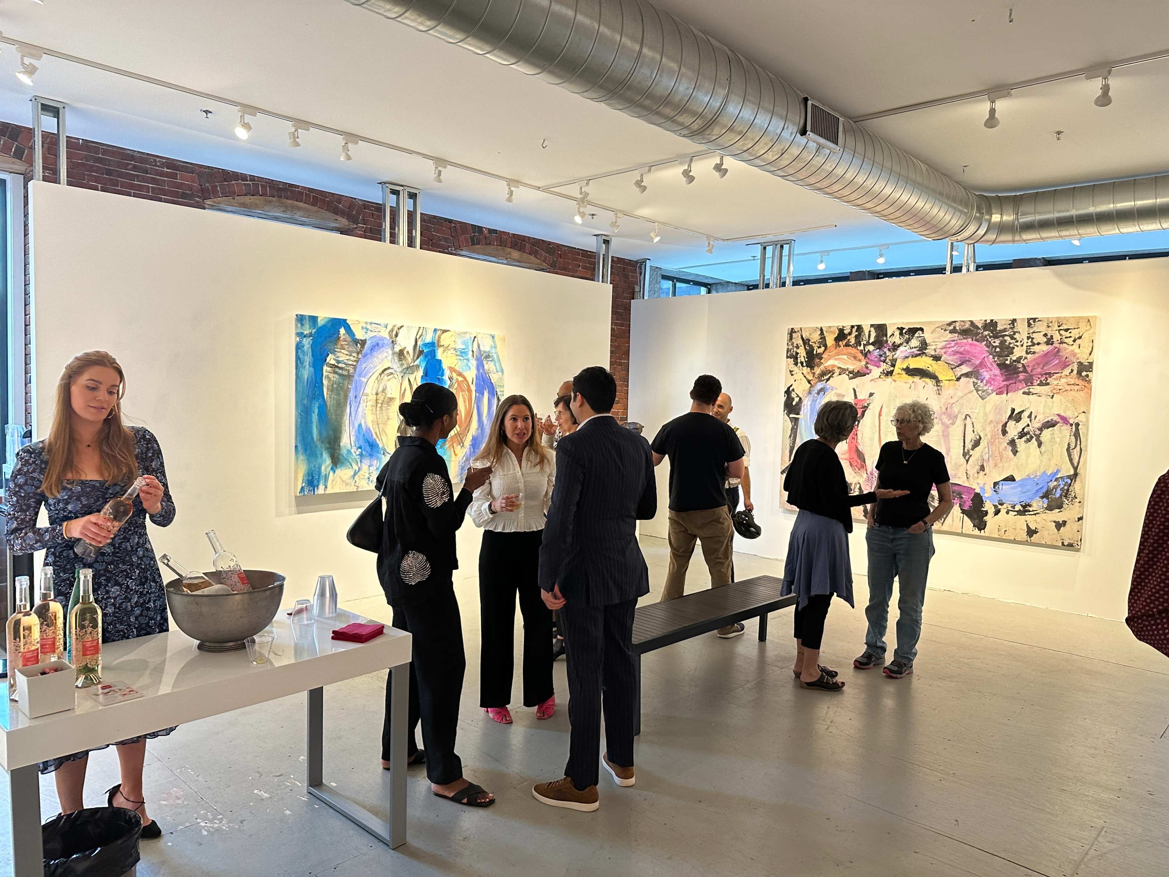 A group of people engages in conversation at an art gallery featuring colorful abstract paintings on the walls.