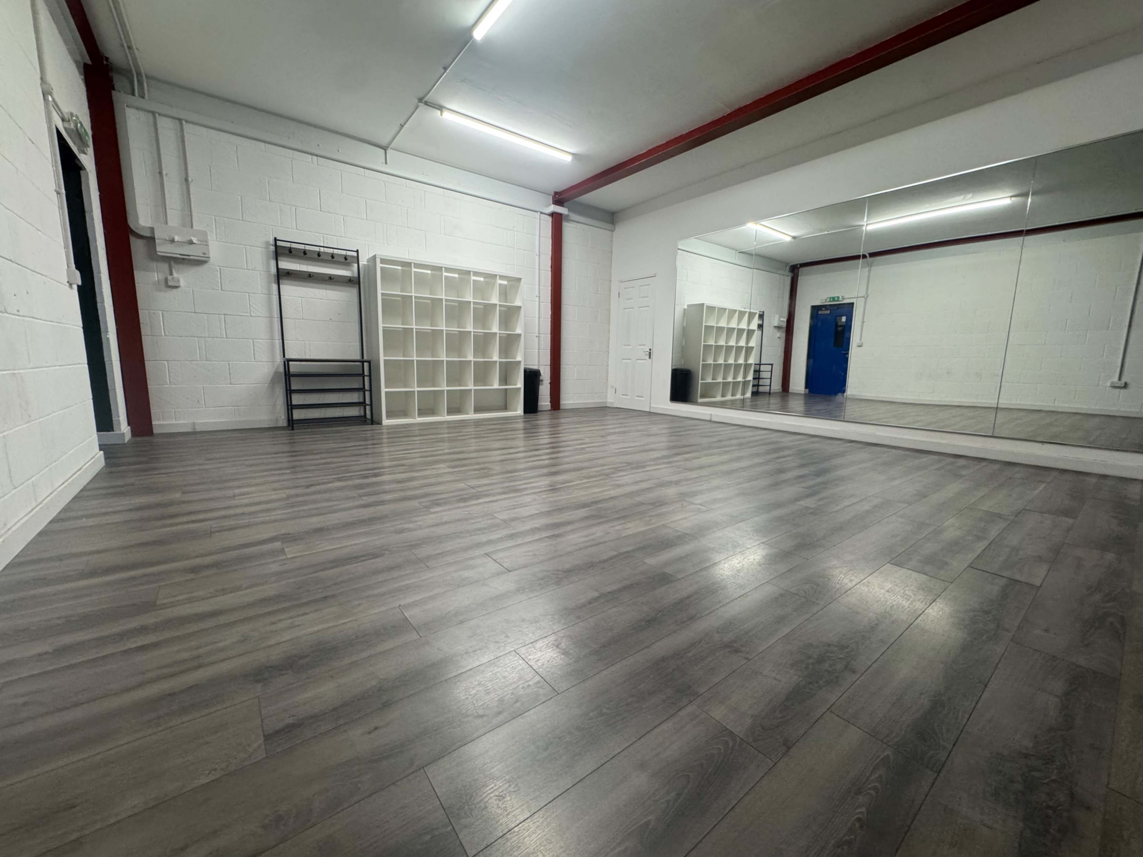 The image shows a spacious room with wooden flooring, white walls, a large mirror, and a shelf unit against one wall.
