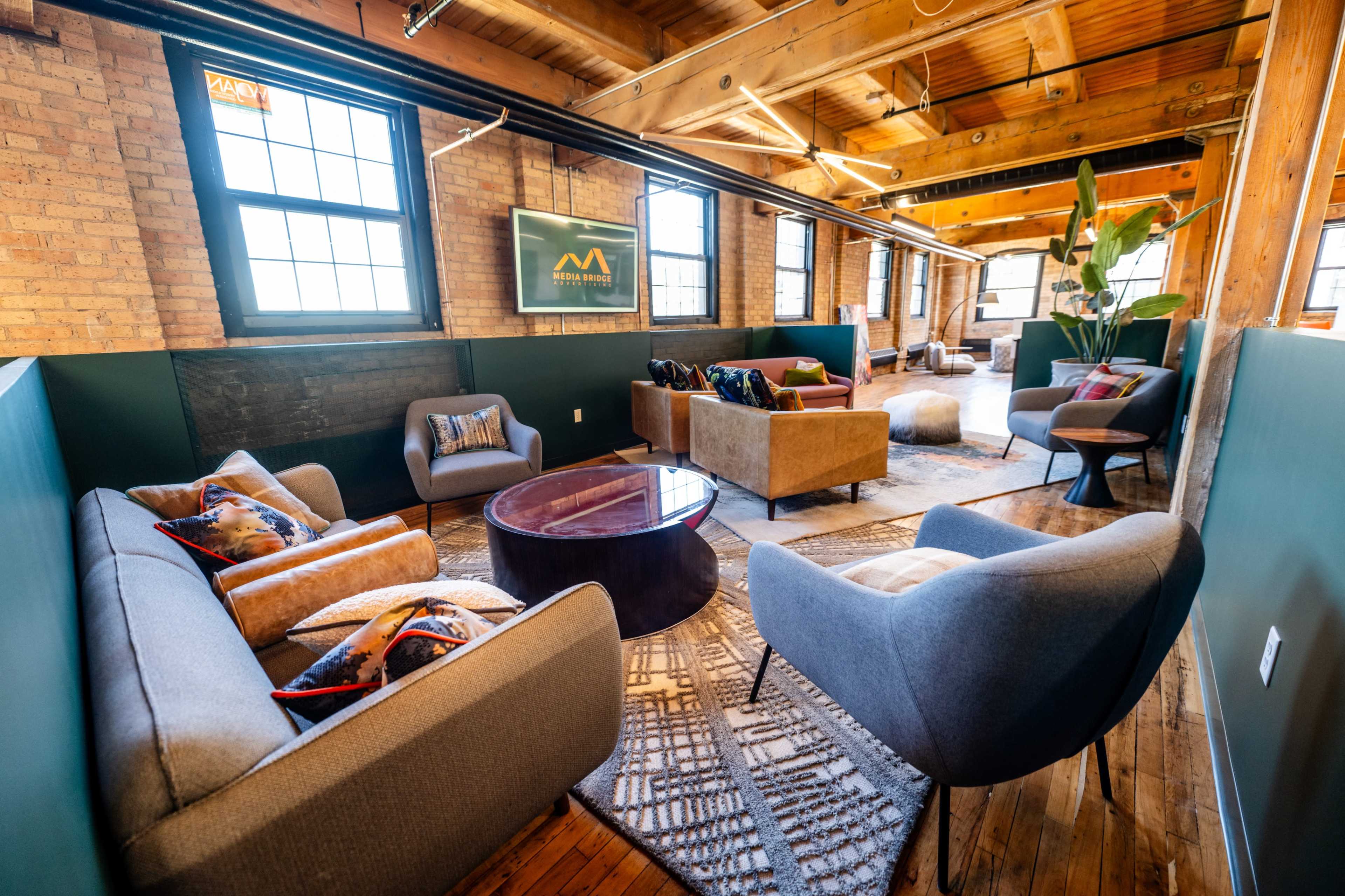 The image shows a modern lounge area with comfortable seating, wooden beams, and large windows, featuring a mix of upholstered chairs and sofas arranged around a circular coffee table.