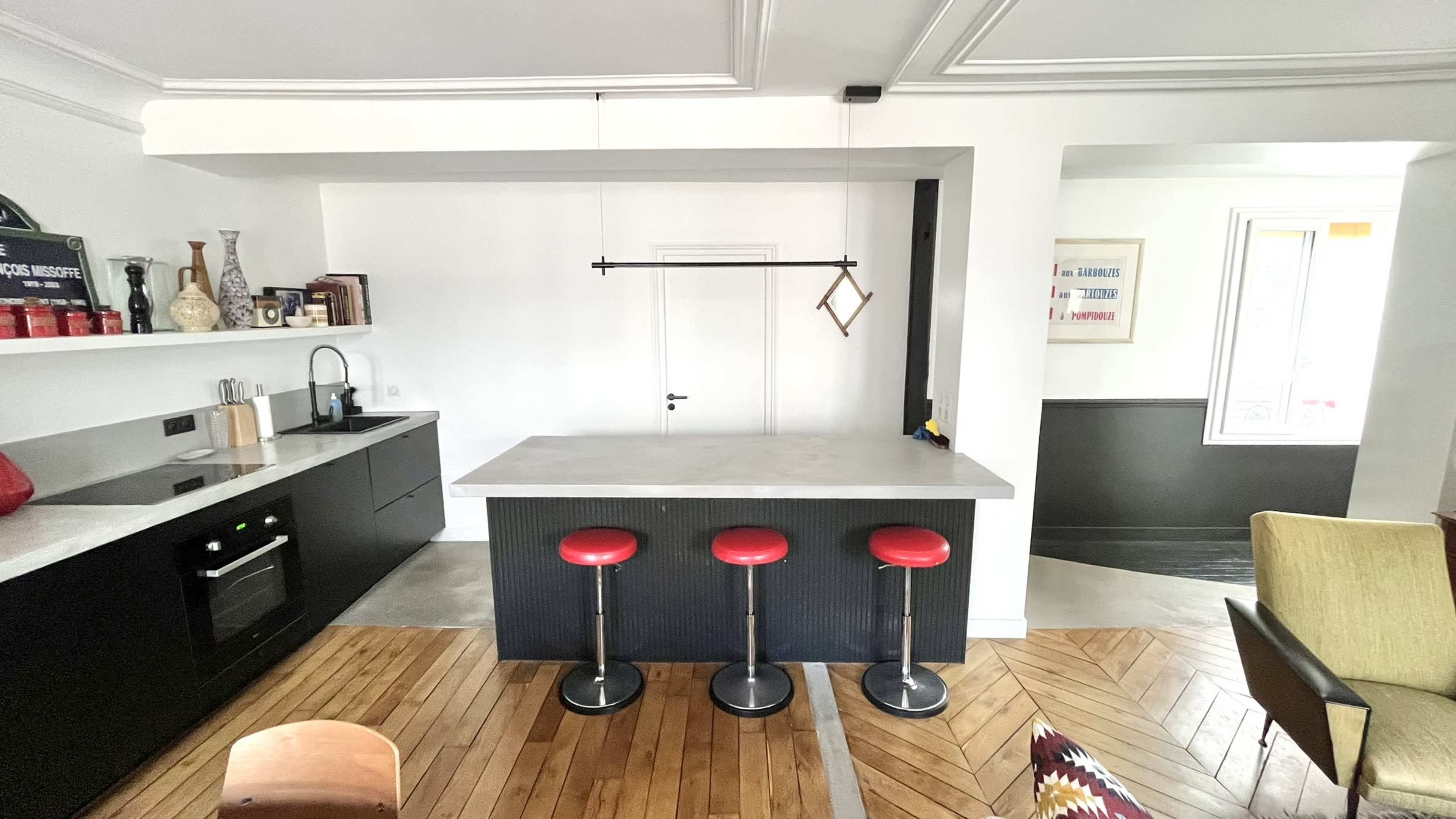 The image depicts a modern kitchen with a minimalist design, featuring a concrete countertop island with three red stools, black cabinetry, and a small dining area with a wooden floor.