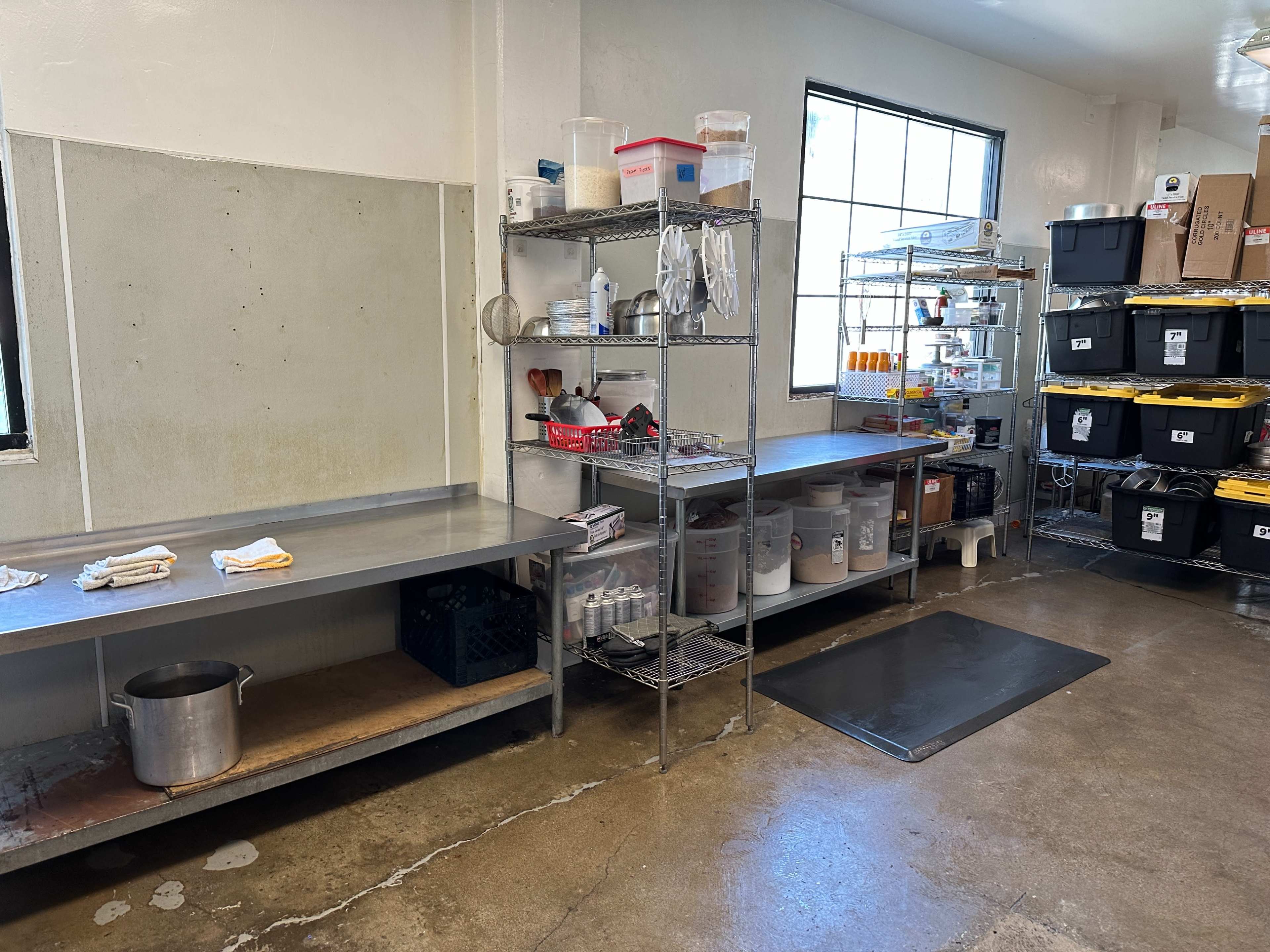 A commercial kitchen area with stainless steel tables, storage shelves containing various containers, and a large pot on the floor.
