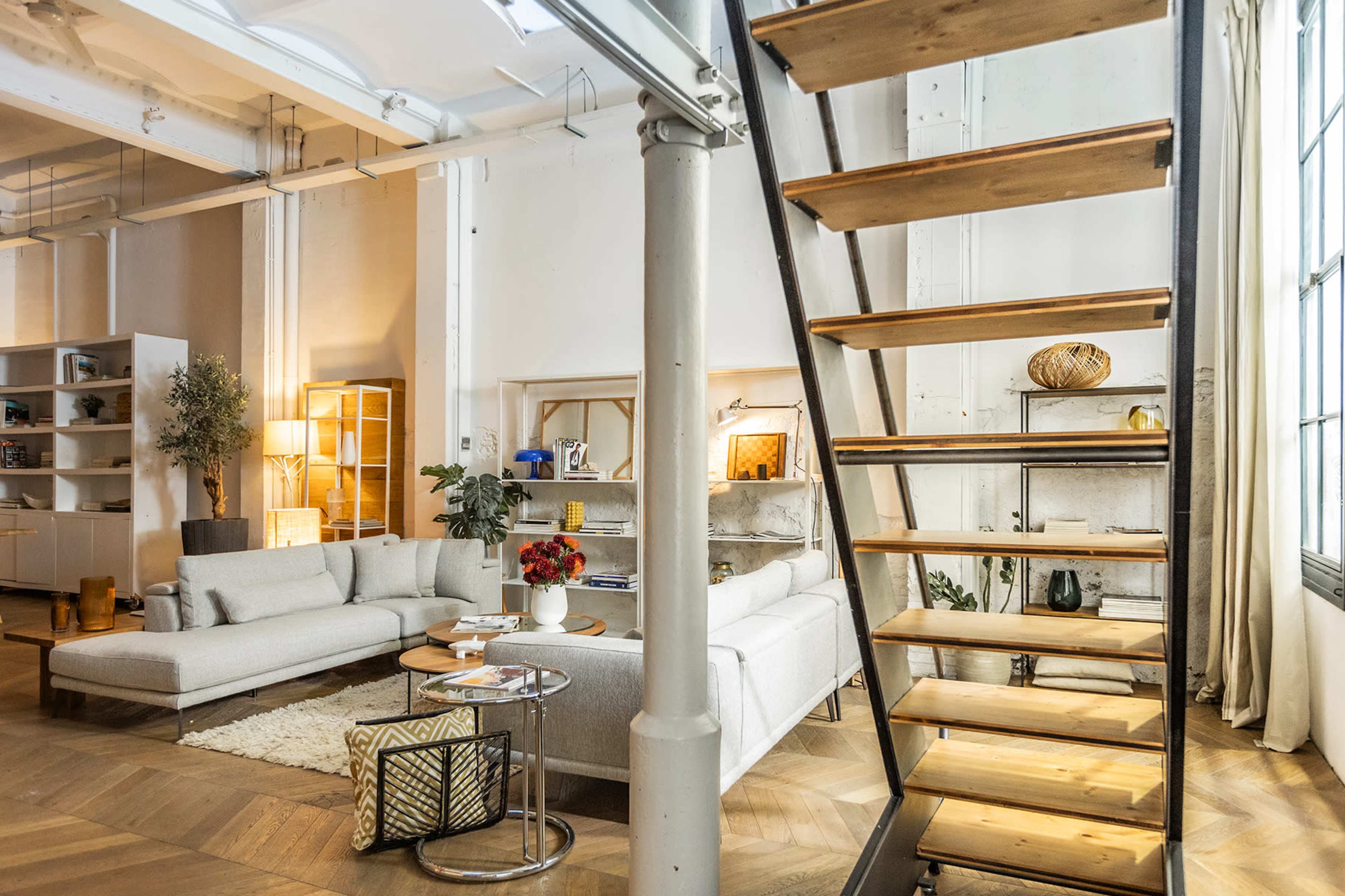 The image shows a modern, open-concept living space featuring a comfortable sofa, a coffee table, a decorative plant, and a wooden staircase leading to an upper level.