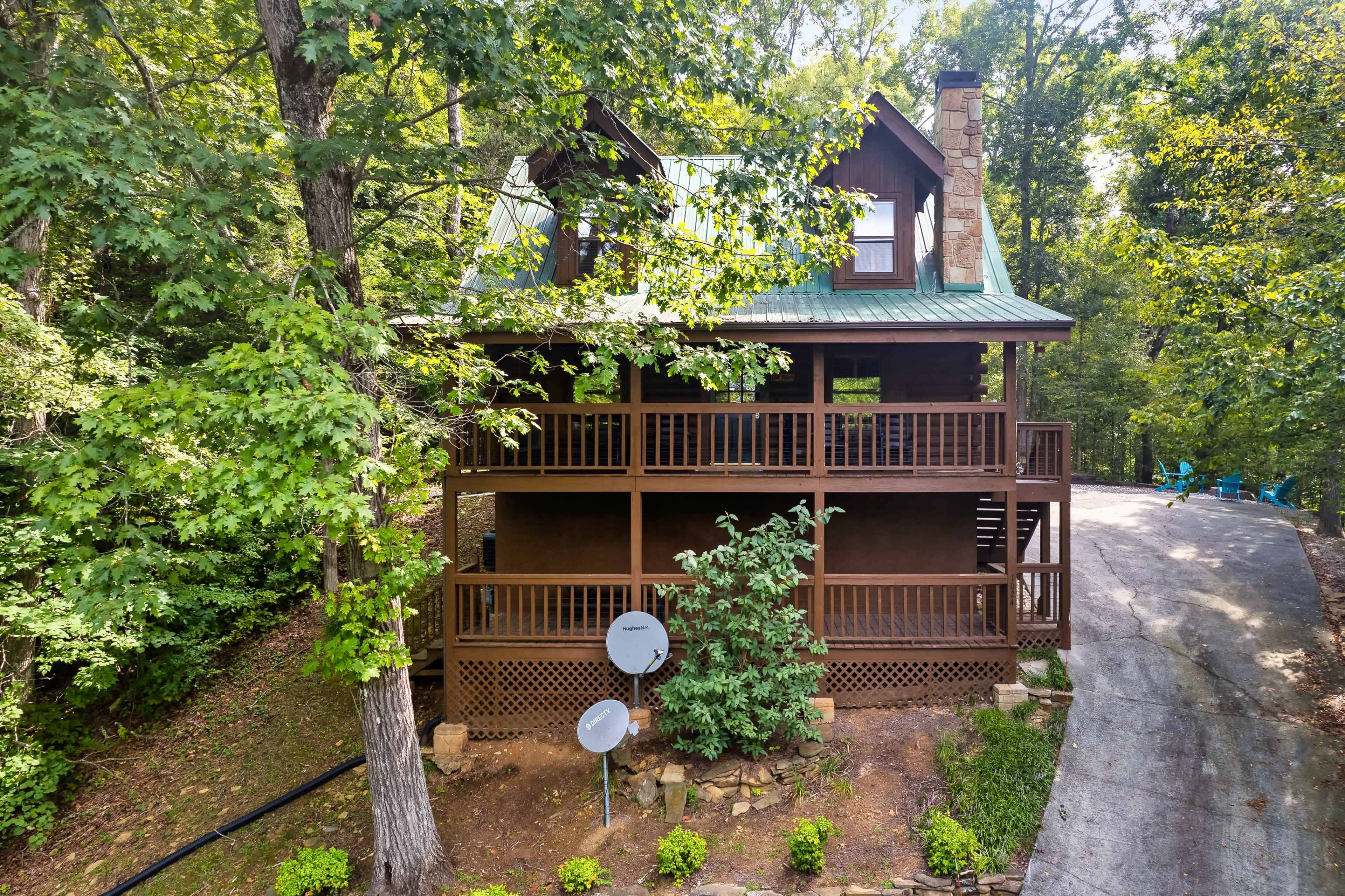 A two-story log cabin with a green metal roof, situated amid trees, with a sloped driveway and satellite dish.