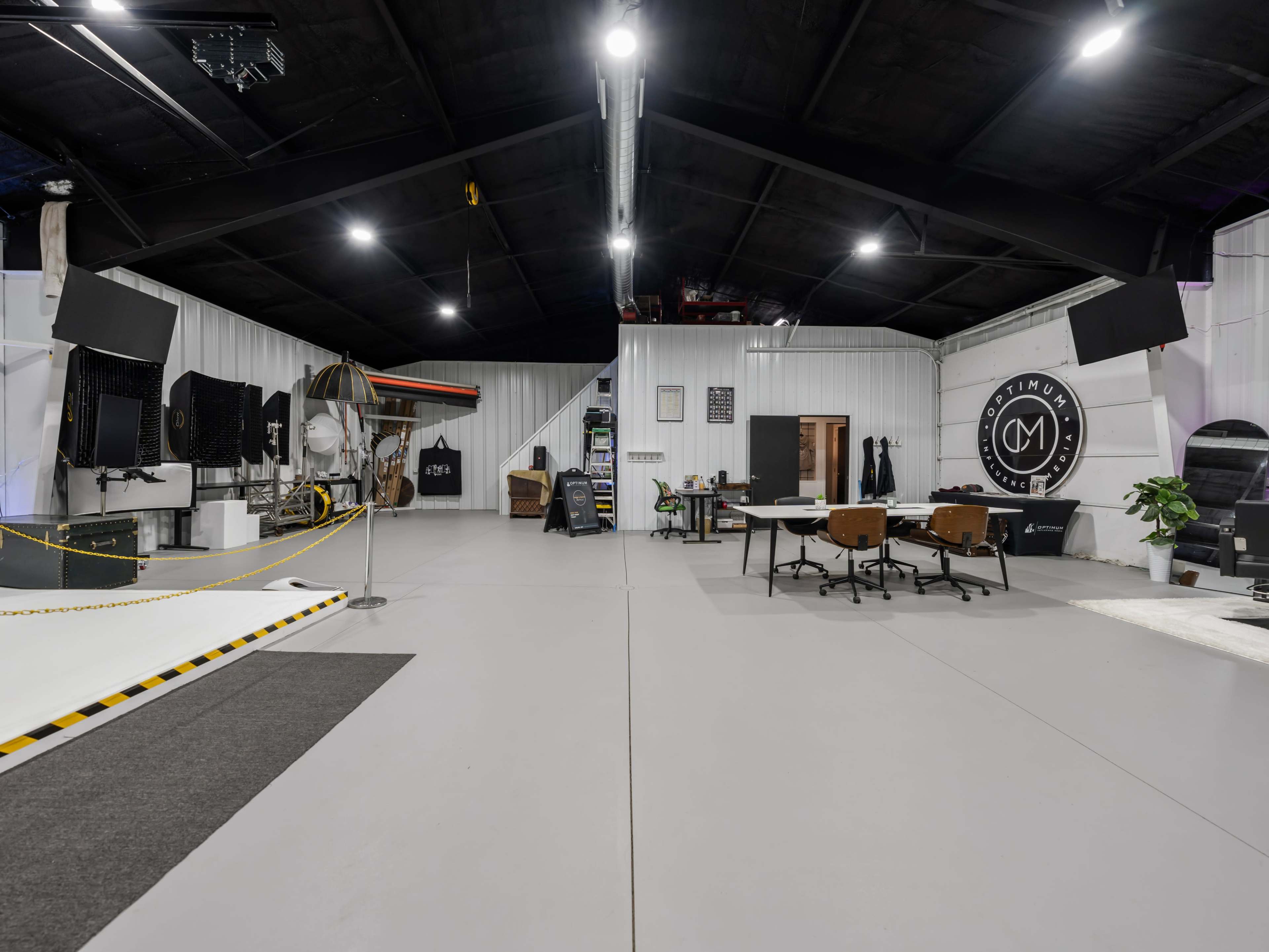 A spacious interior of a modern studio or workspace, featuring a large open area with desks, chairs, and various equipment against a backdrop of white walls and a black ceiling.