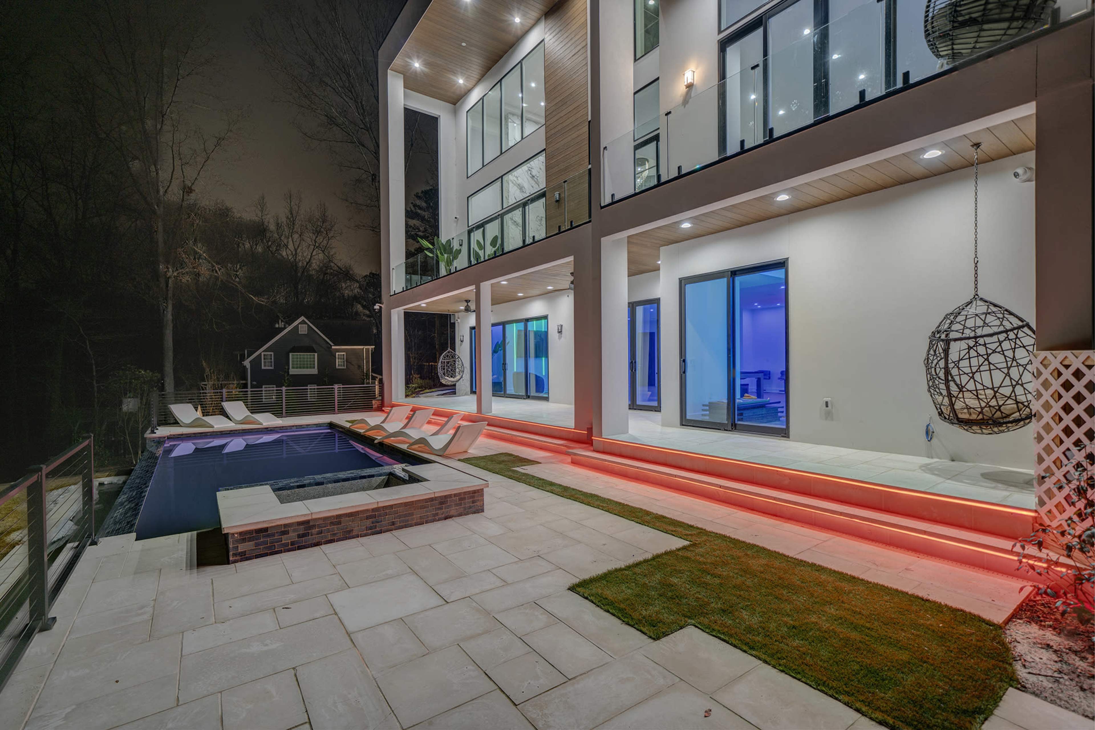 A modern two-story house features a sleek pool area with illuminated steps, a hanging chair, and large windows showcasing a brightly lit interior.