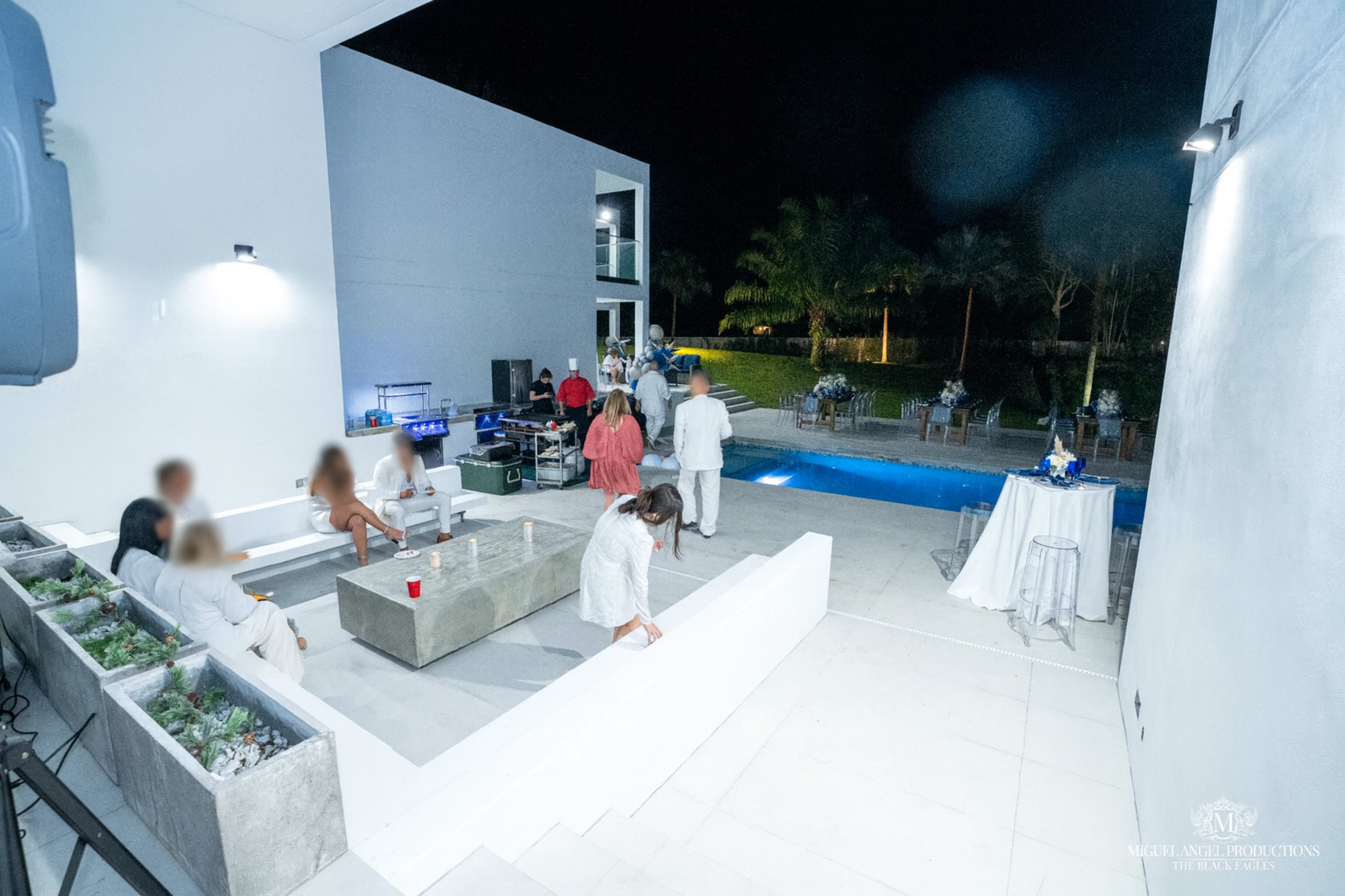 A group of people is gathered around a pool in a modern outdoor space at night, with some guests seated and others standing near a bar area.