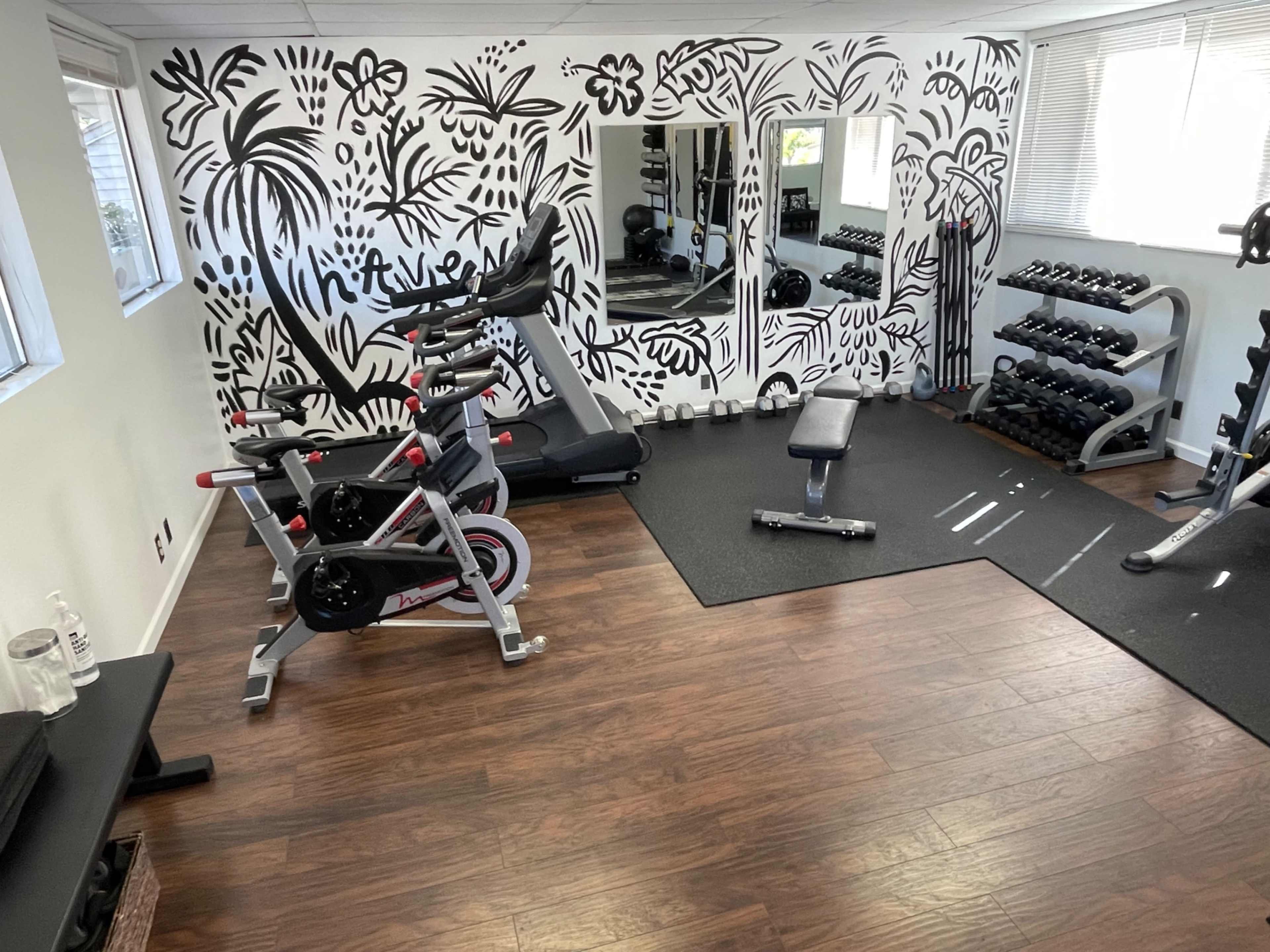 The image shows a home gym with exercise equipment, including a stationary bike, a bench, and a rack of dumbbells, set against a patterned black-and-white mural.
