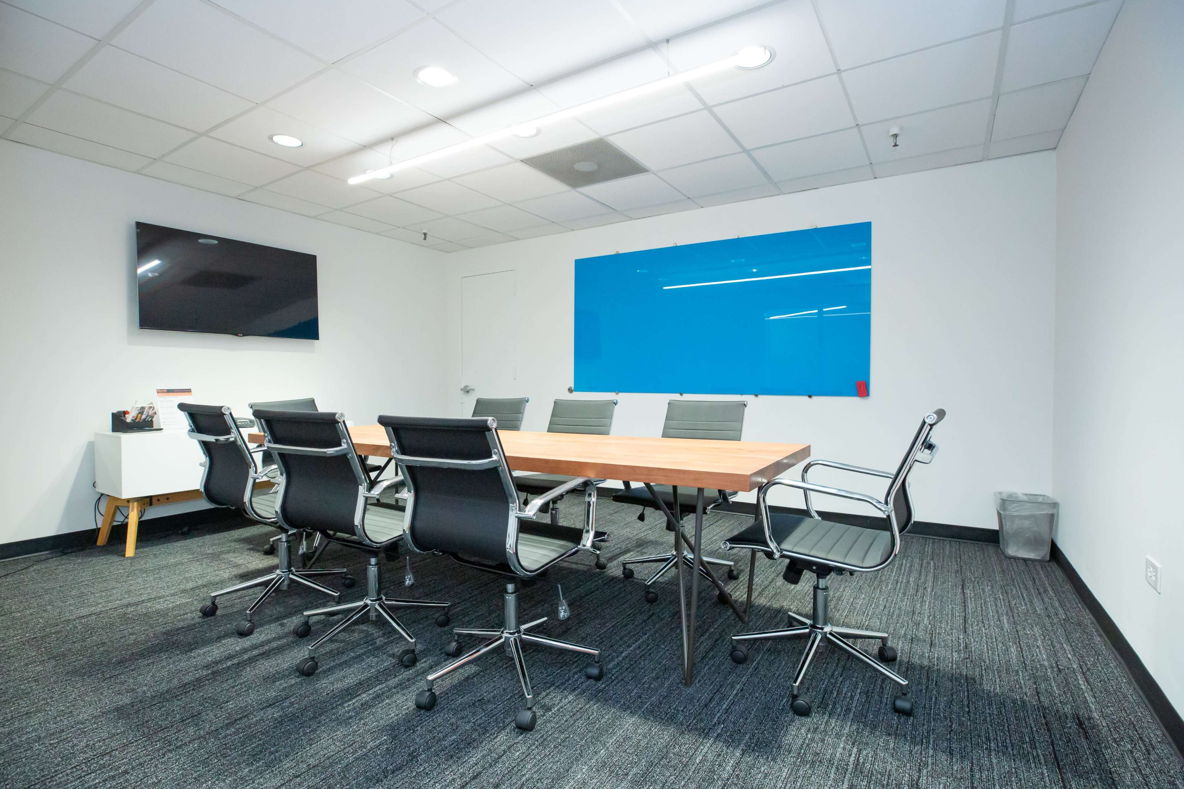 The image shows a modern conference room featuring a long wooden table surrounded by black office chairs, with a television on one wall and a blue whiteboard on another.