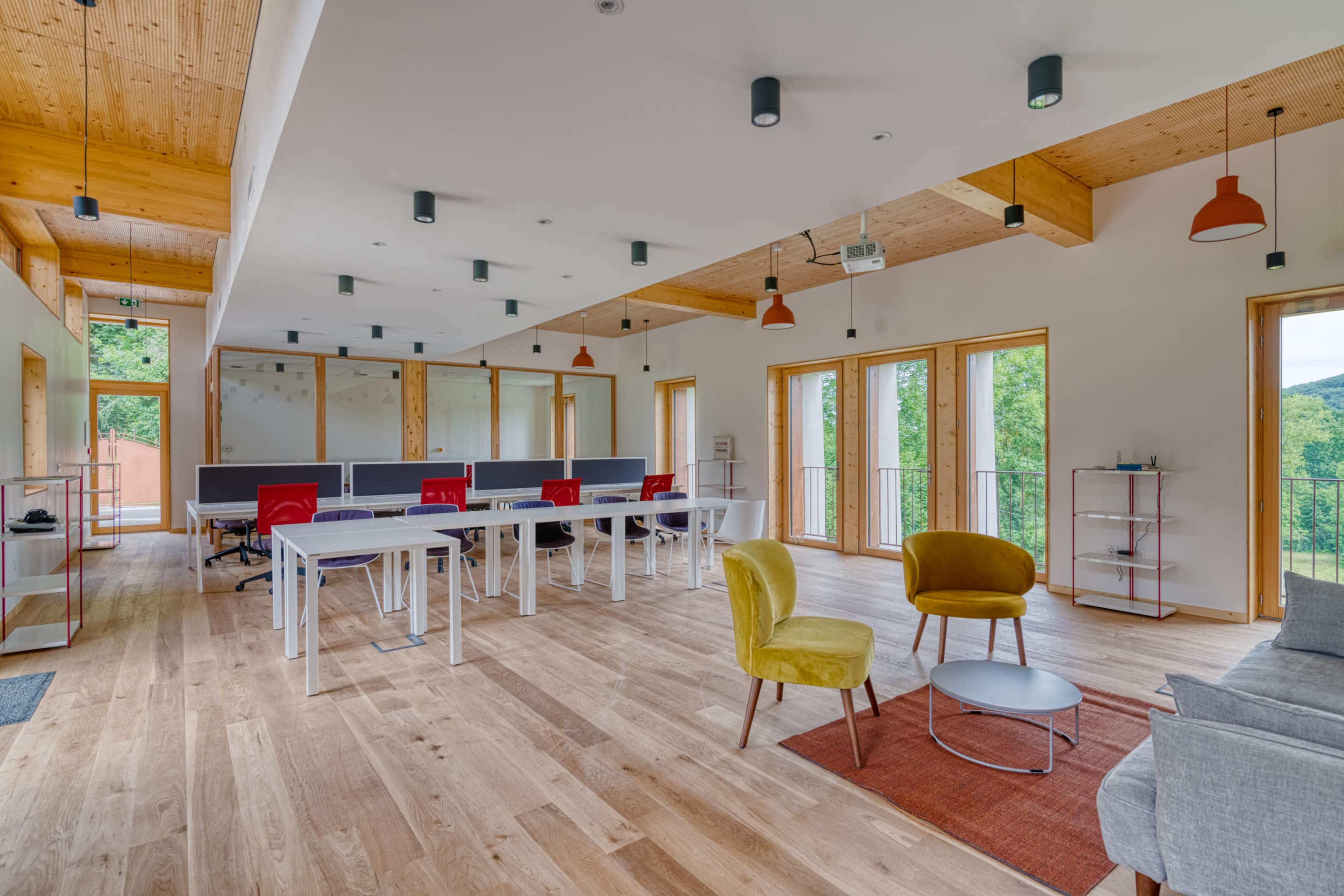 A modern open office space features a row of desks, colorful chairs, and large windows that provide natural light and a view of greenery outside.