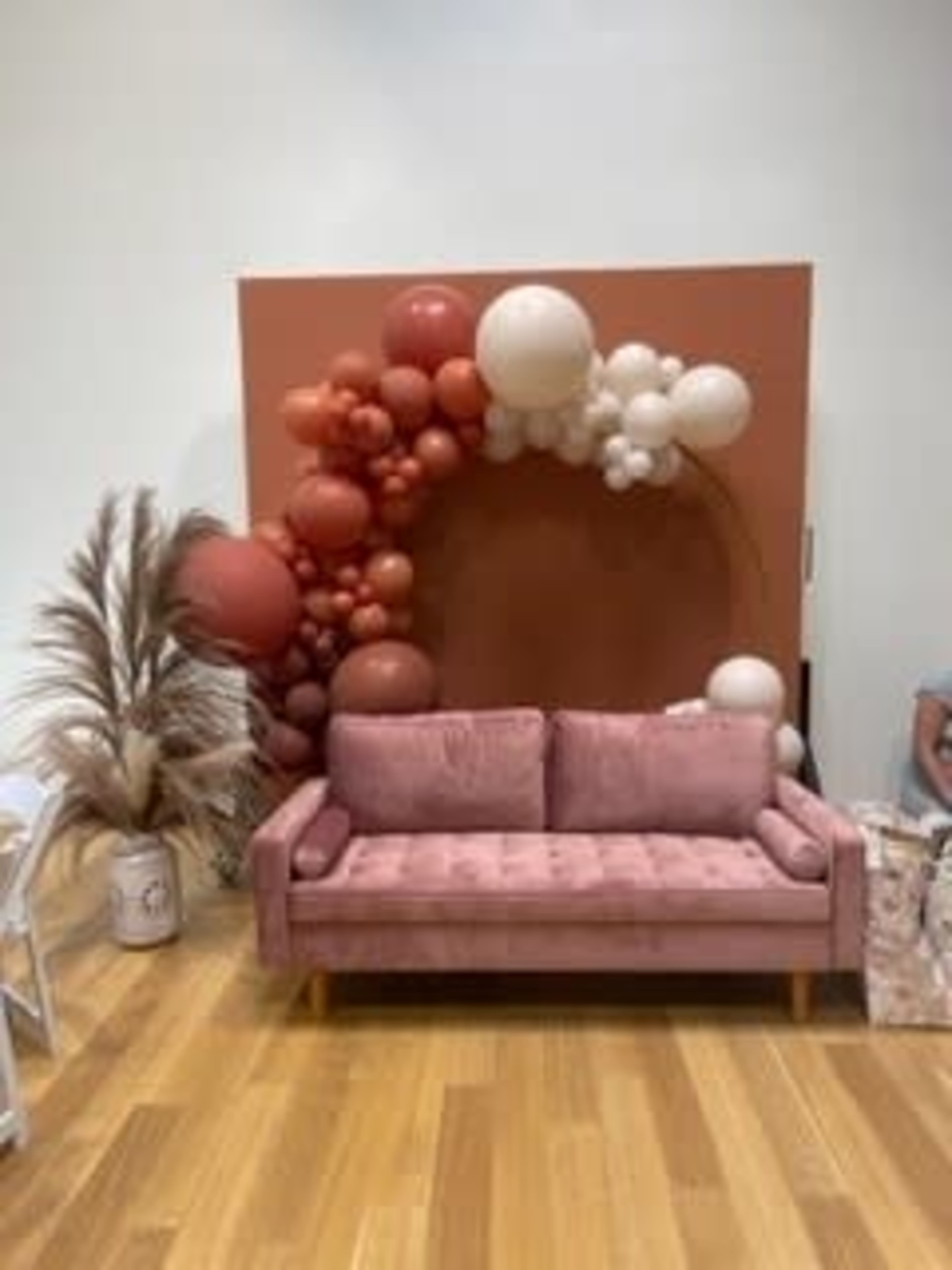 A pink sofa in front of a decorative wall adorned with an arrangement of balloons and dried palm leaves.