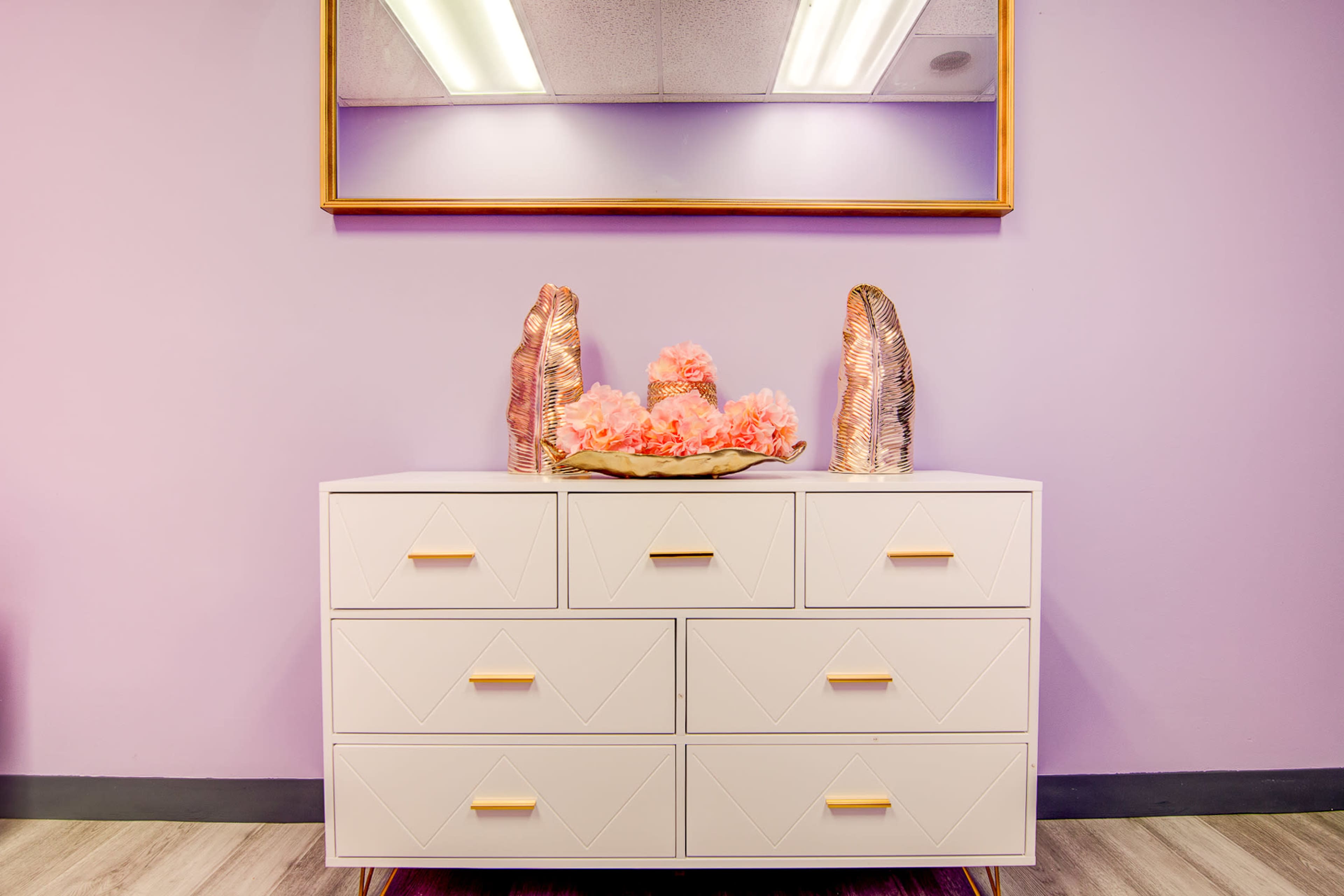 A white dresser with six drawers, topped with a decorative tray holding pink flowers and flanked by two textured sculptures, against a purple wall and a mirror.