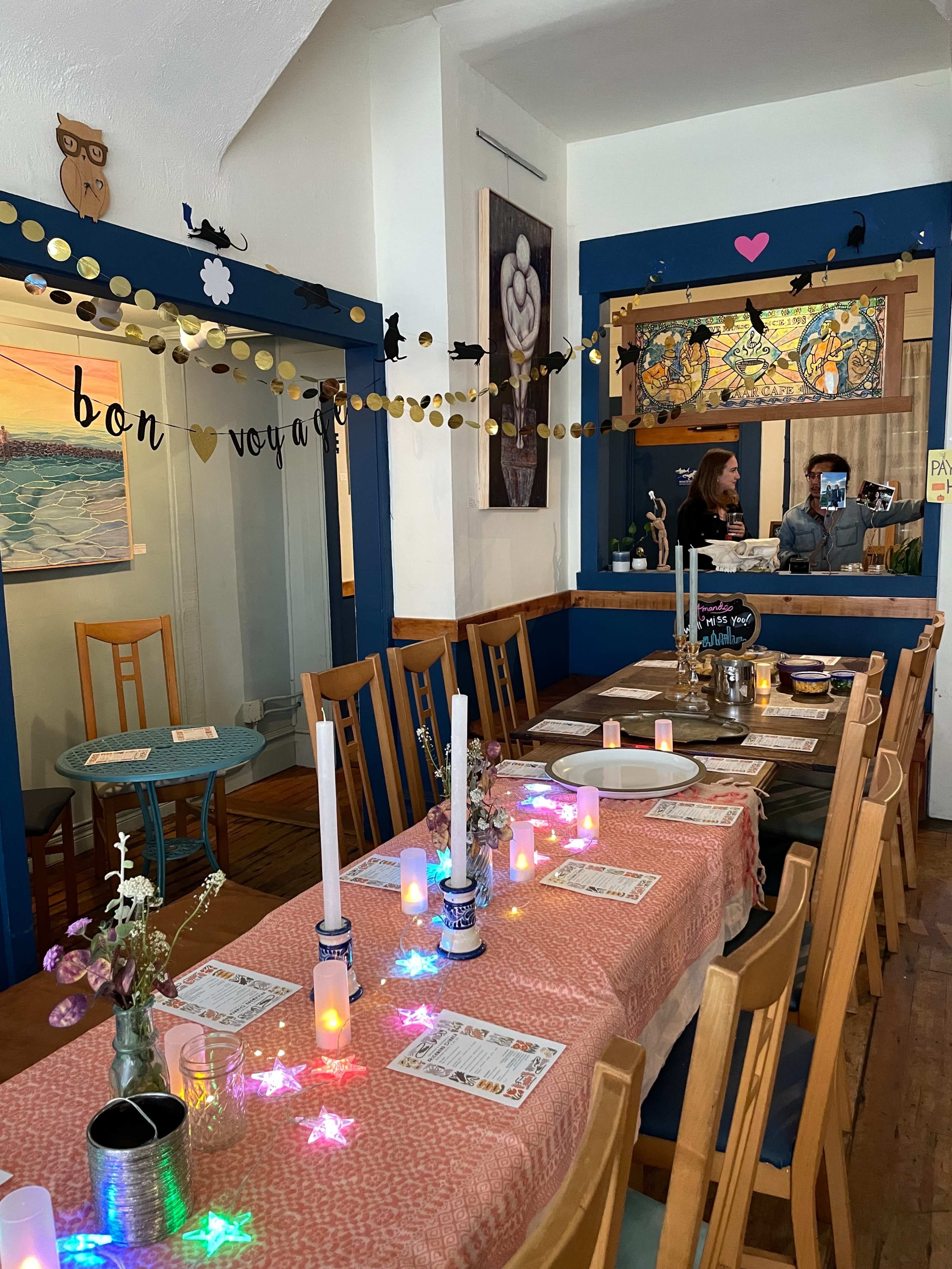 A cozy dining area with a decorated table set for a meal, featuring candles, flowers, and colorful lights, alongside a cheerful banner that reads "bon voyage."