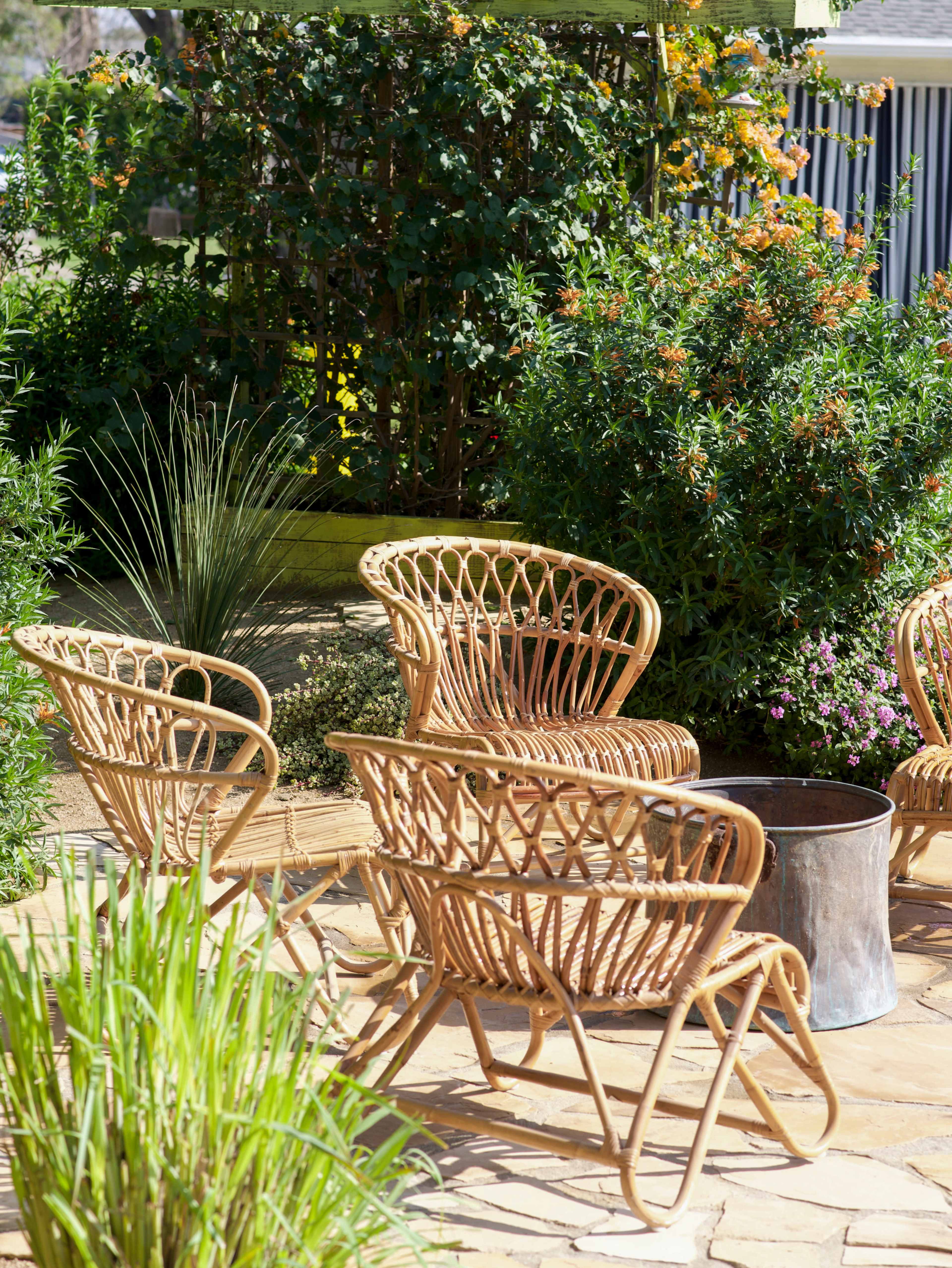 The image shows a patio area with four rattan chairs arranged around a small metal table, surrounded by green plants and shrubs.