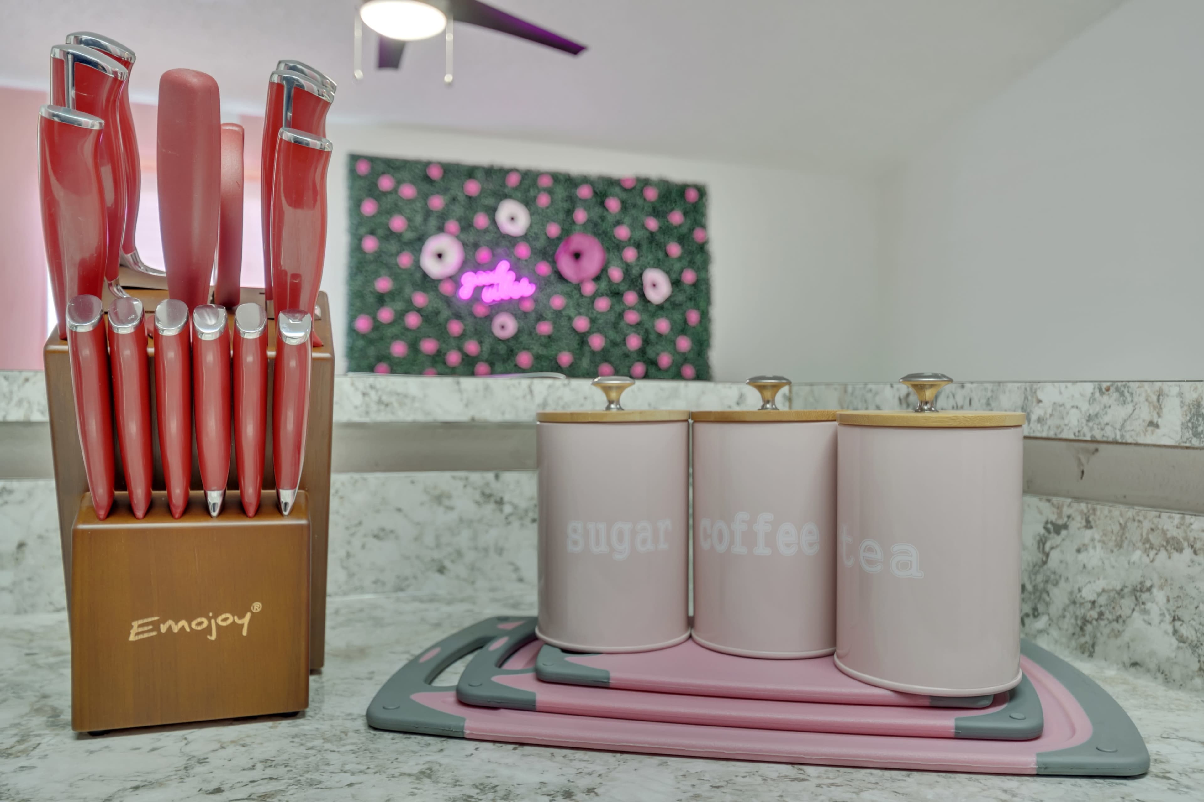 A wooden knife block holds pink knives beside three pink canisters labeled "sugar," "coffee," and "tea" on a counter.