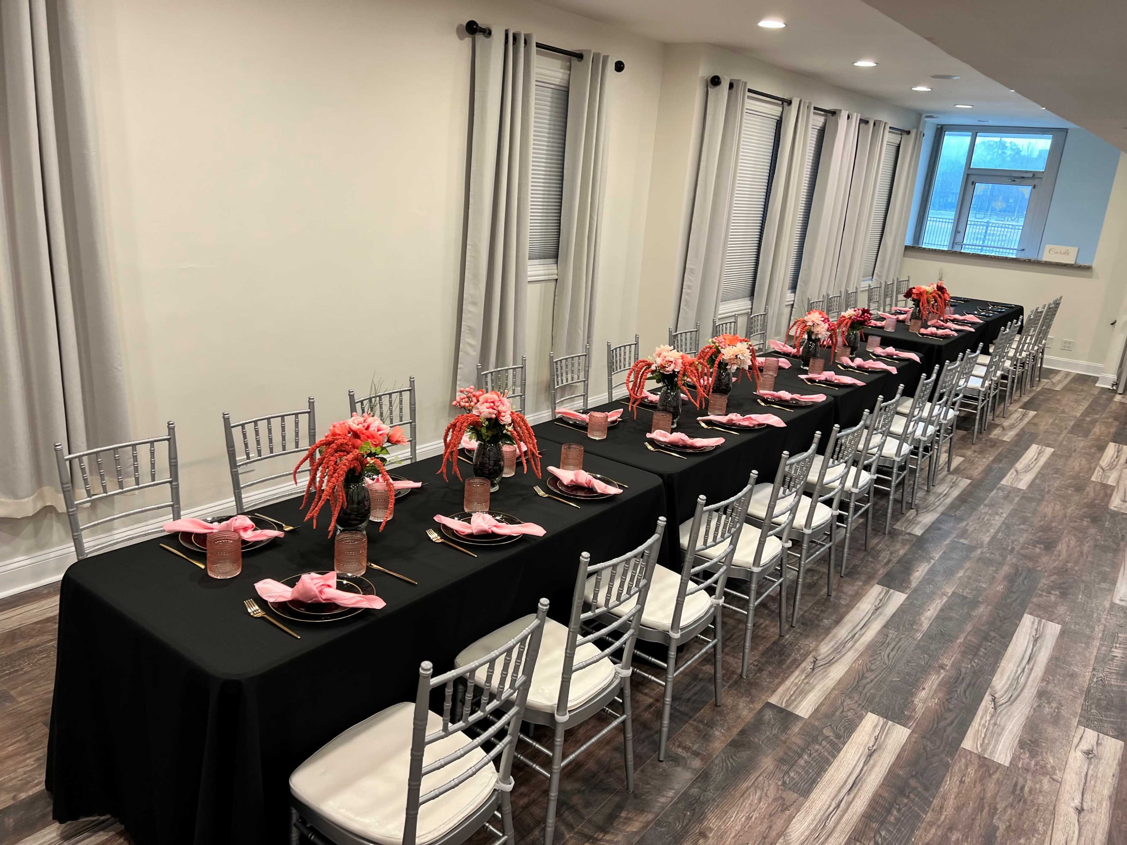 A long banquet table is set with black tablecloths, pink napkins, and floral centerpieces, lined with silver chiavari chairs in a well-lit room.