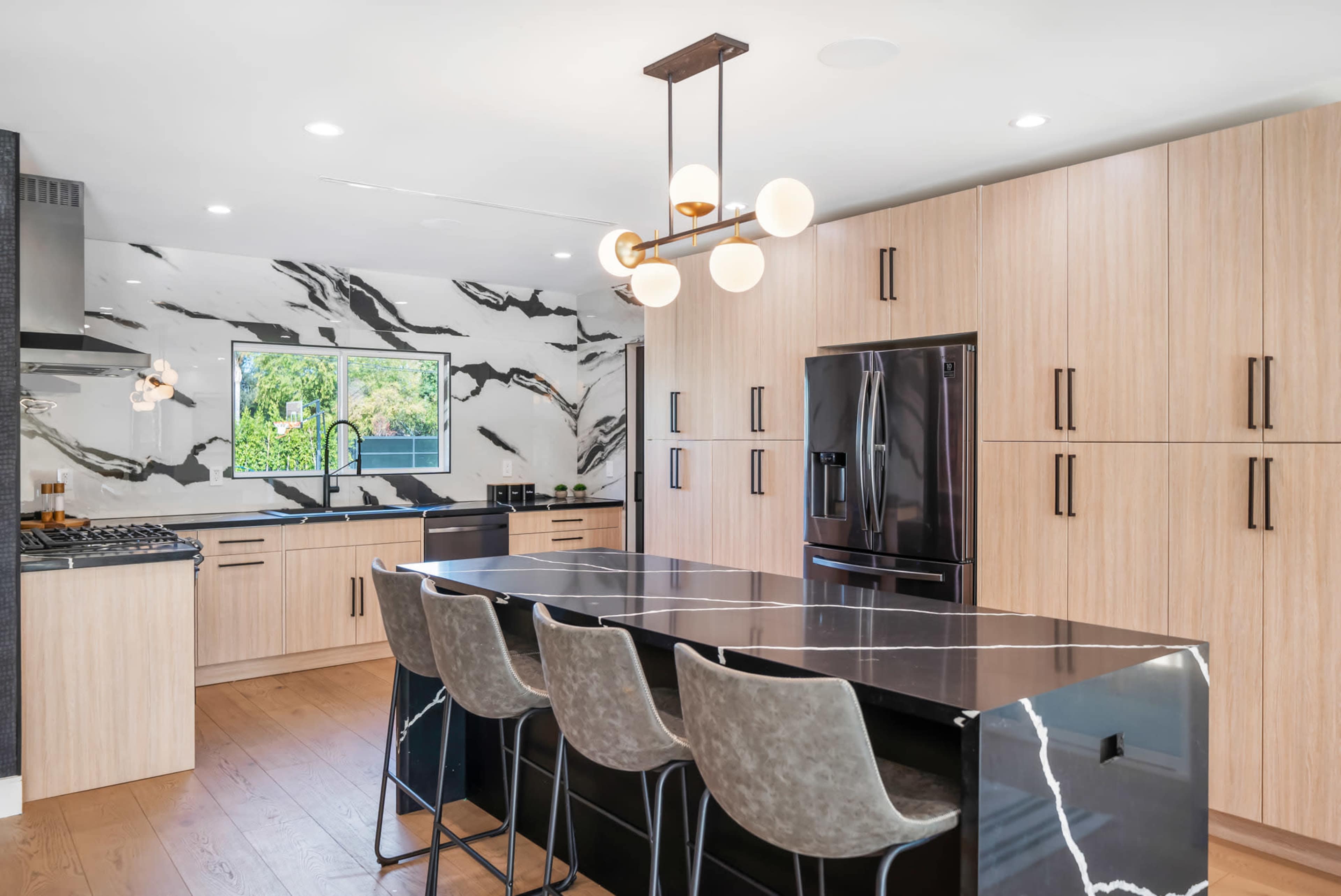 A modern kitchen featuring a large island with black marble countertops, light wood cabinetry, and a striking marble backsplash.