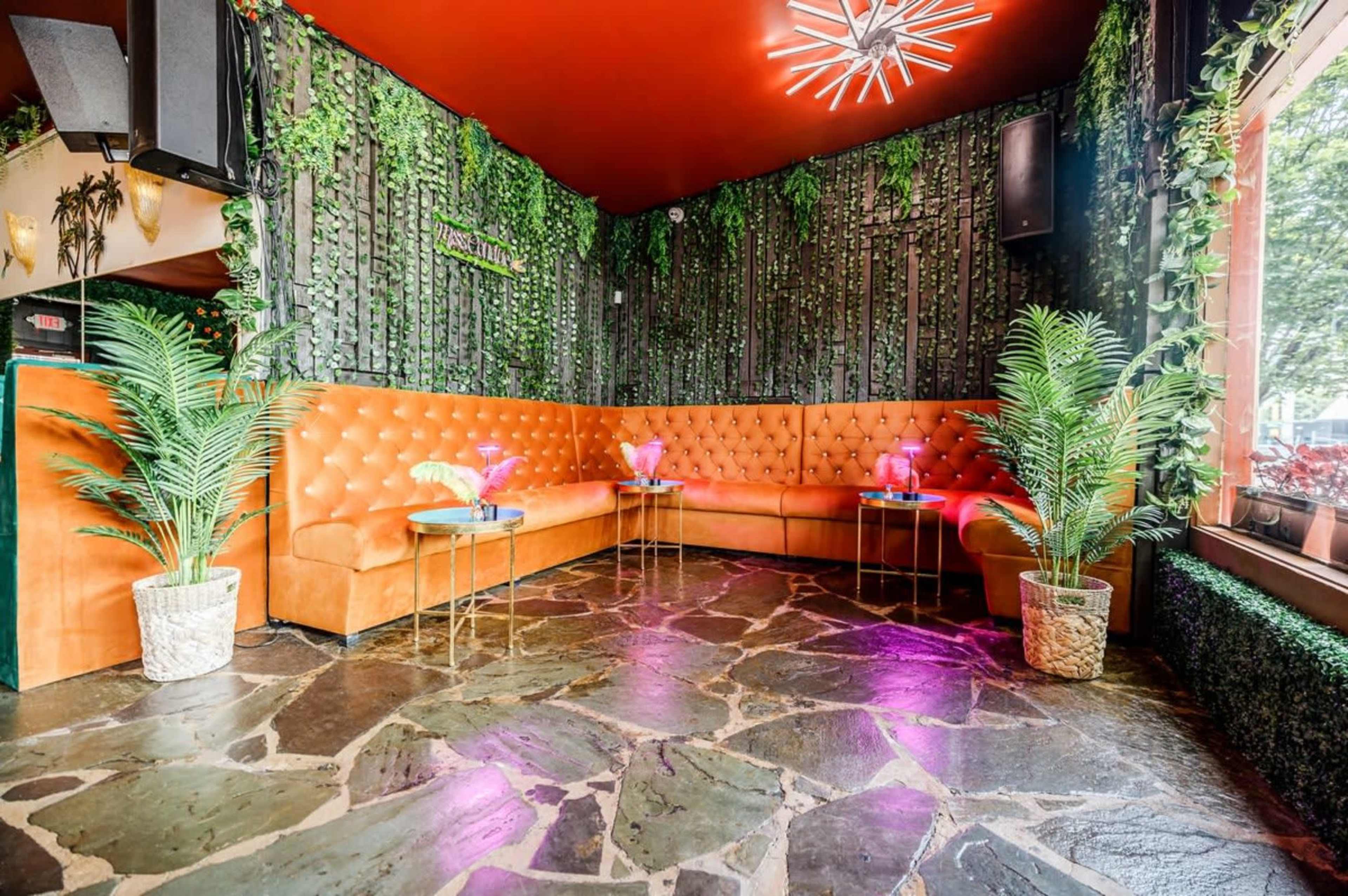 The image shows a cozy lounge area with a large orange tufted couch, green plants, and stone flooring illuminated by pink lighting.