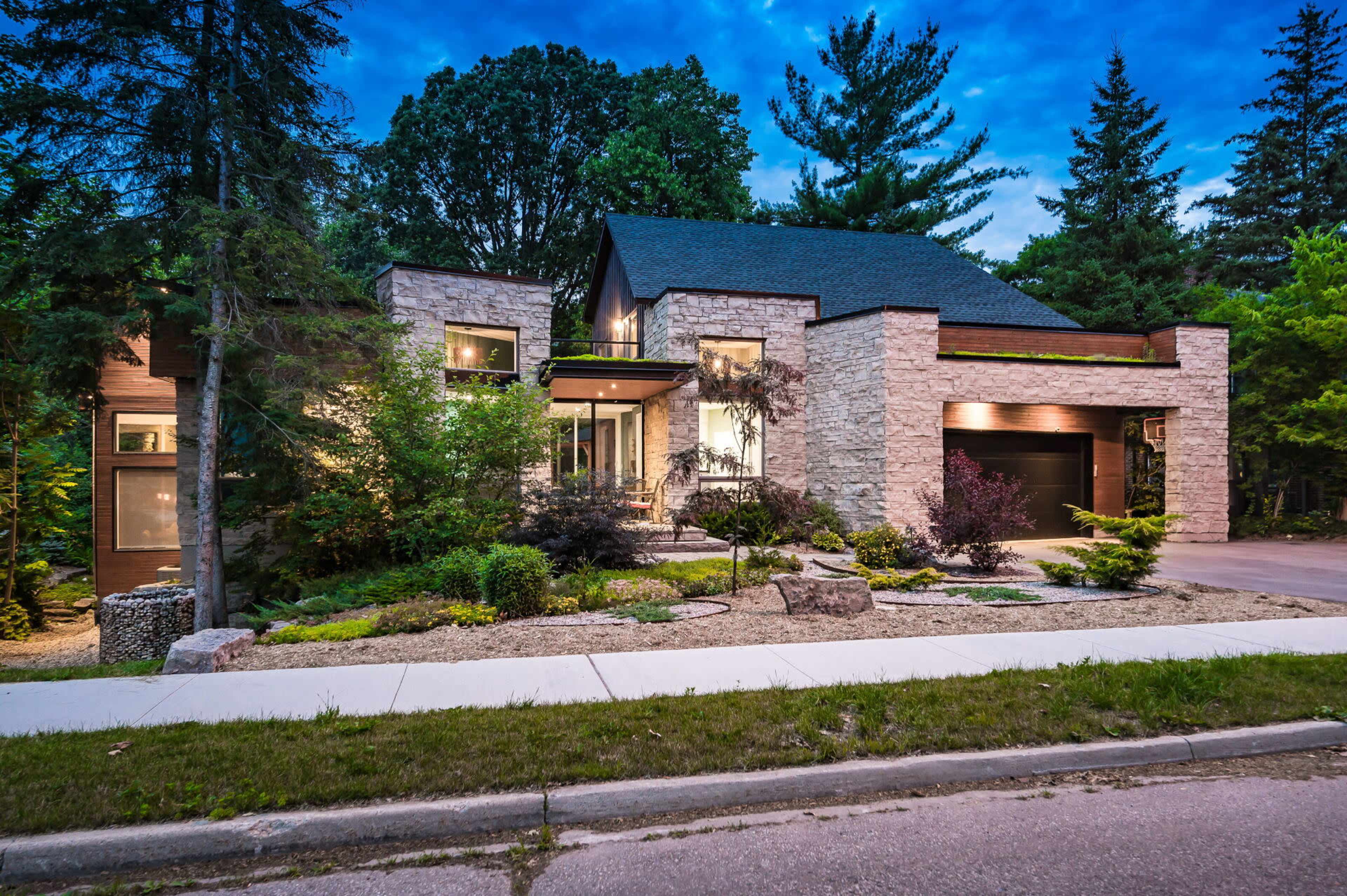 A modern stone and wood house is surrounded by lush greenery and a landscaped yard, with a paved walkway leading to the entrance.