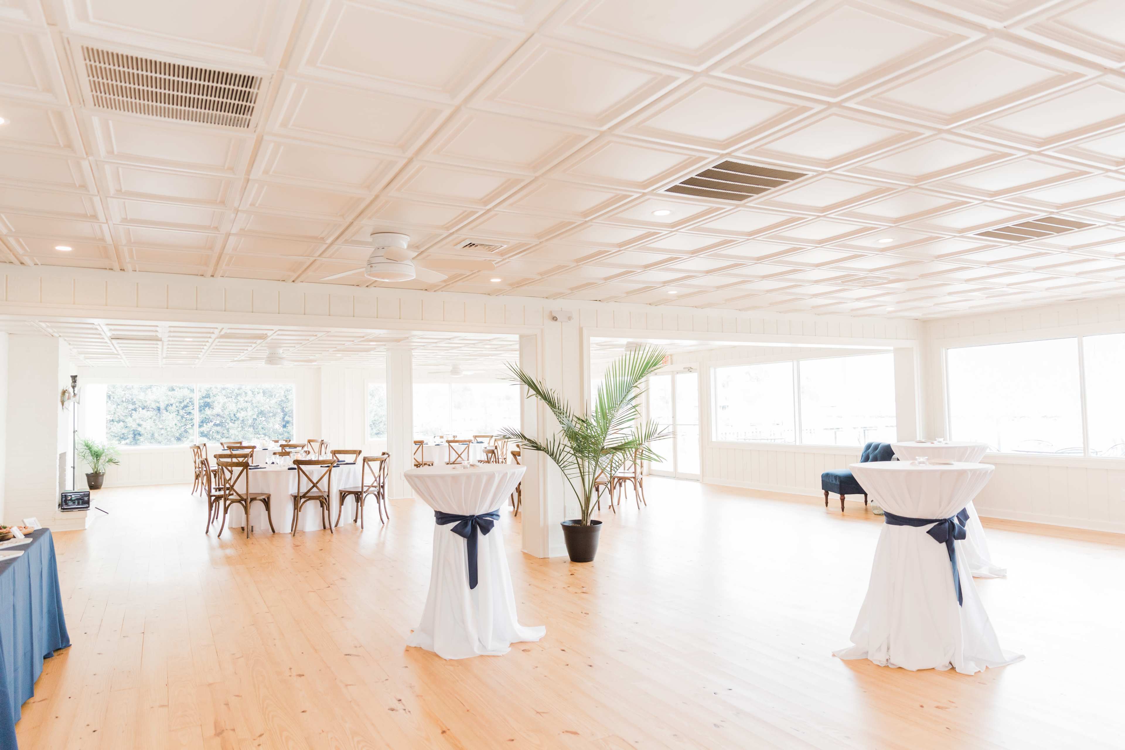 A spacious event room with wooden flooring, a white ceiling, and several round tables set with white tablecloths and navy blue sashes.