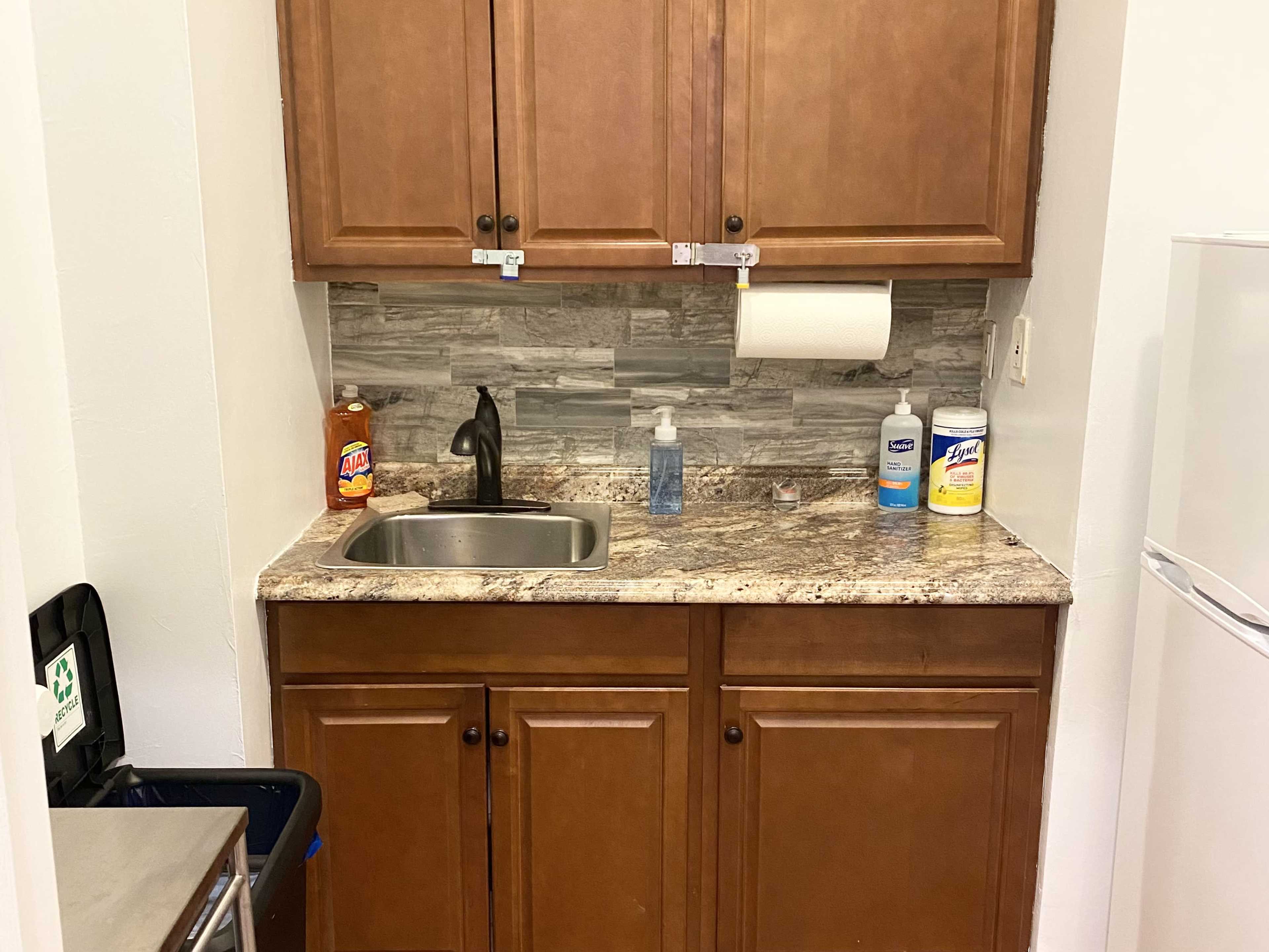 A small kitchen area with a stainless steel sink, a granite countertop, various cleaning supplies, and brown wooden cabinets above.