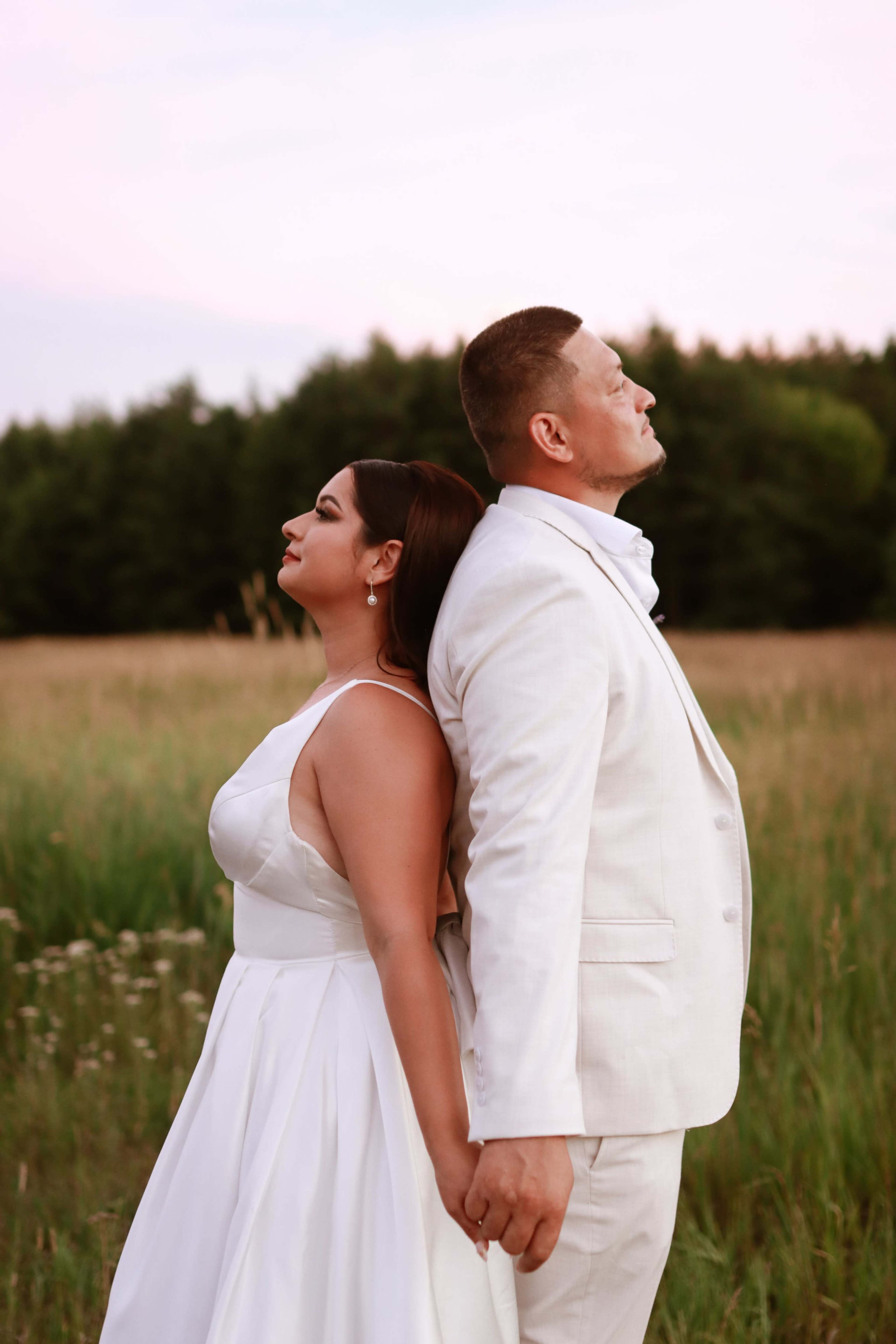 A man and a woman stand back-to-back in a grassy field, holding hands while looking away from each other.