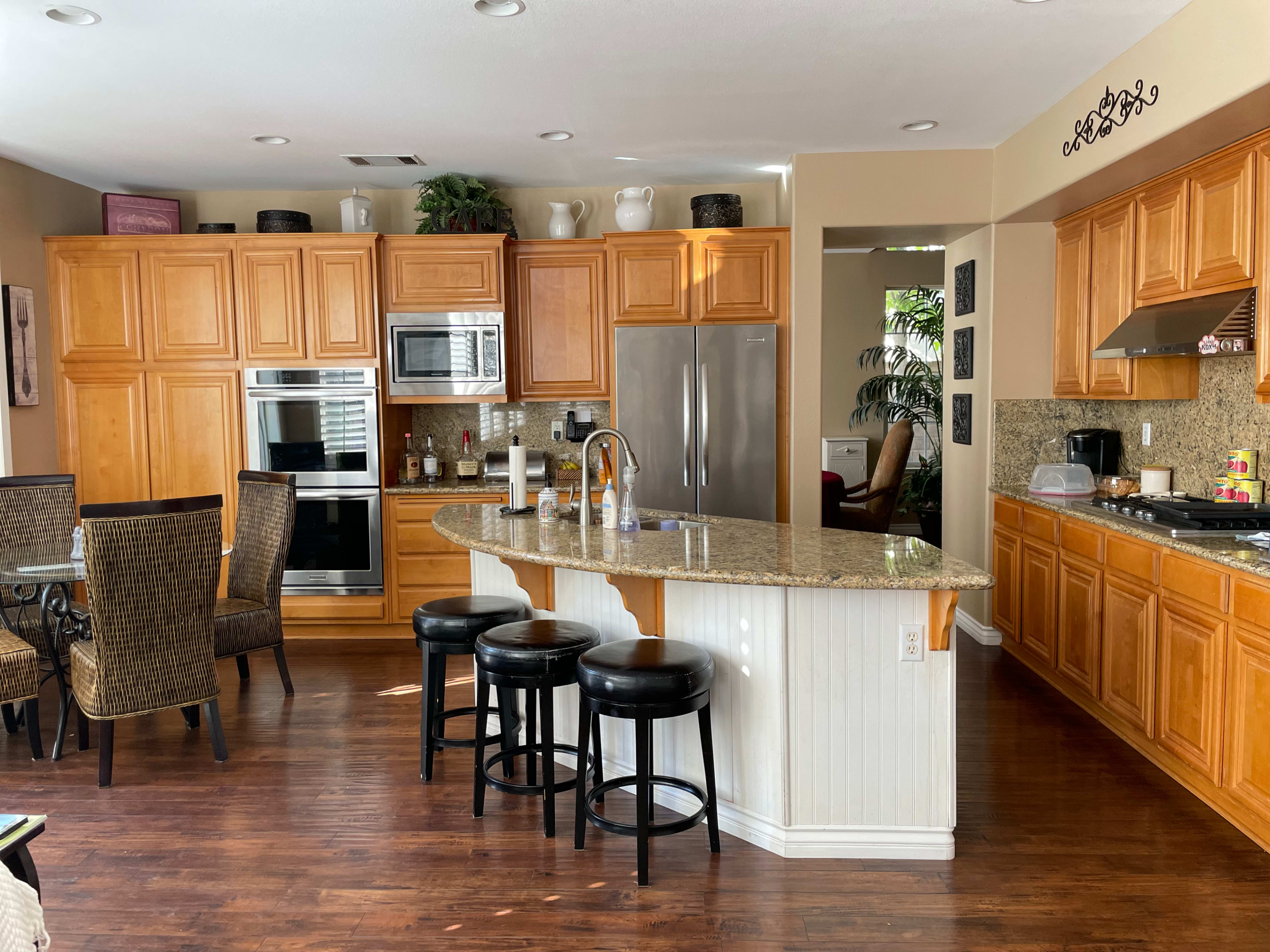 A kitchen with wooden cabinets, a granite countertop island, black bar stools, and a dining area with wicker chairs.