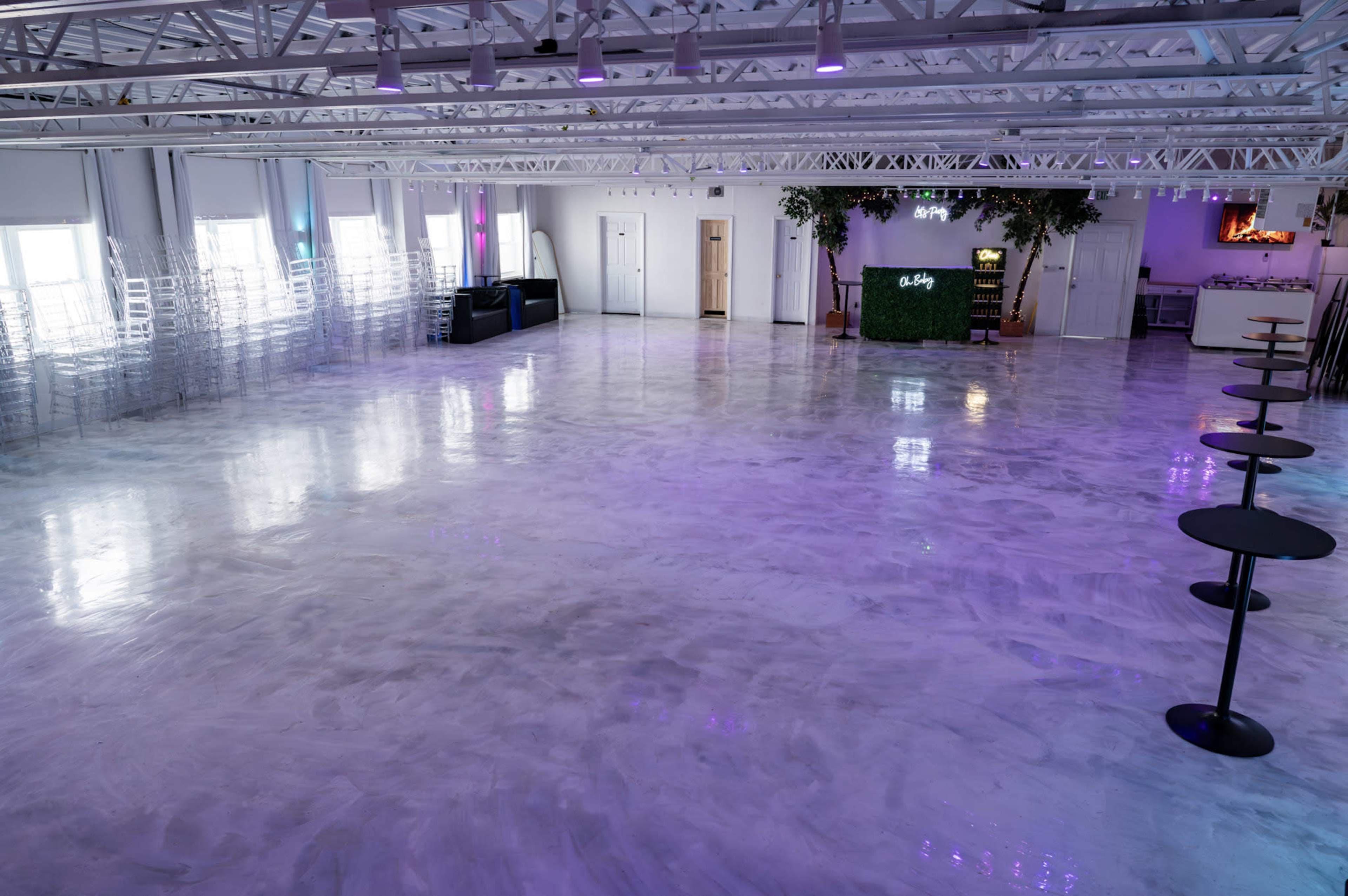 The image shows a spacious event hall with a polished floor, white walls, and several tables arranged along one side.