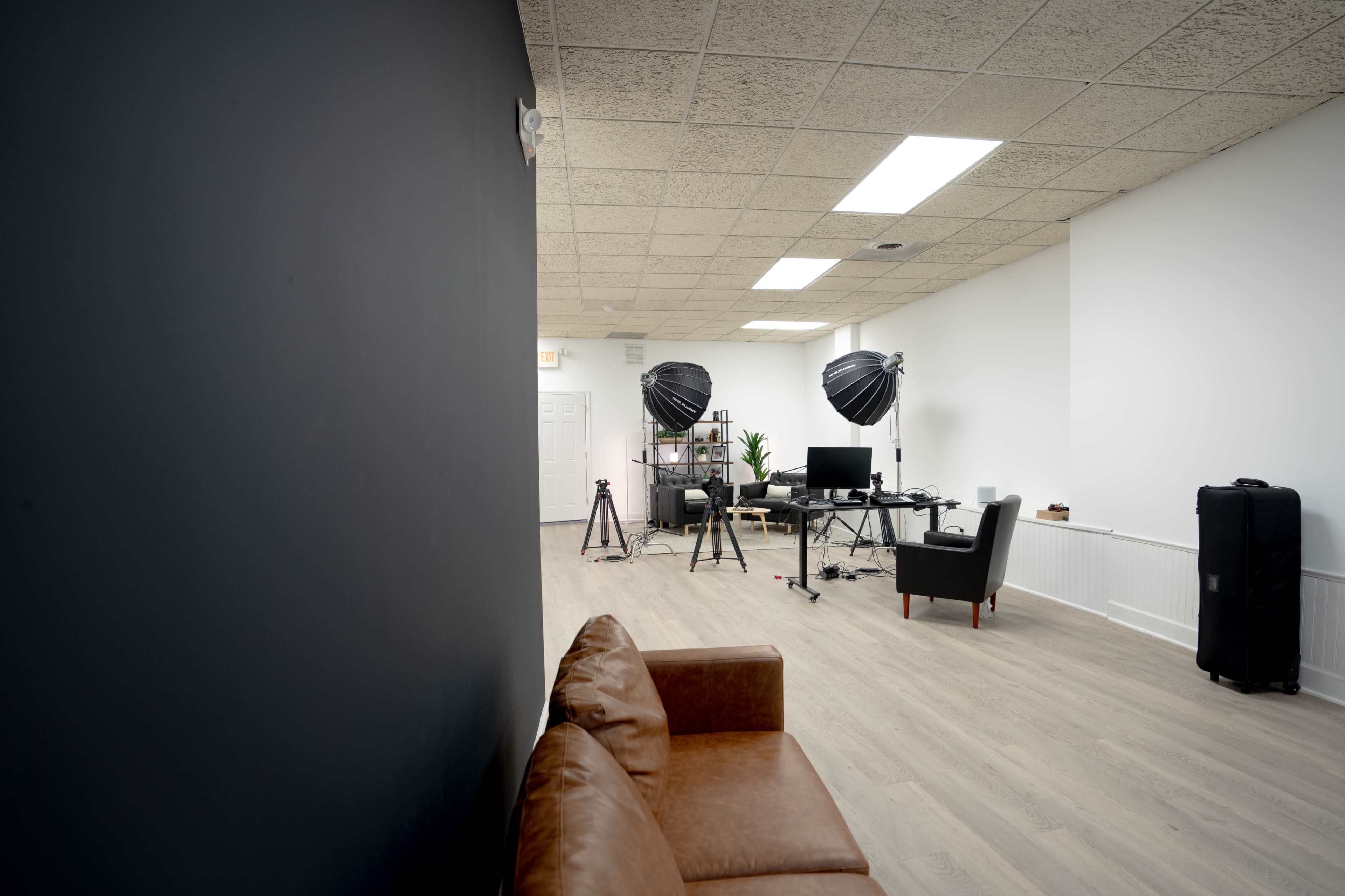 The image shows a spacious interior office with a brown leather couch, desk setup with photography equipment, and large softbox lights.
