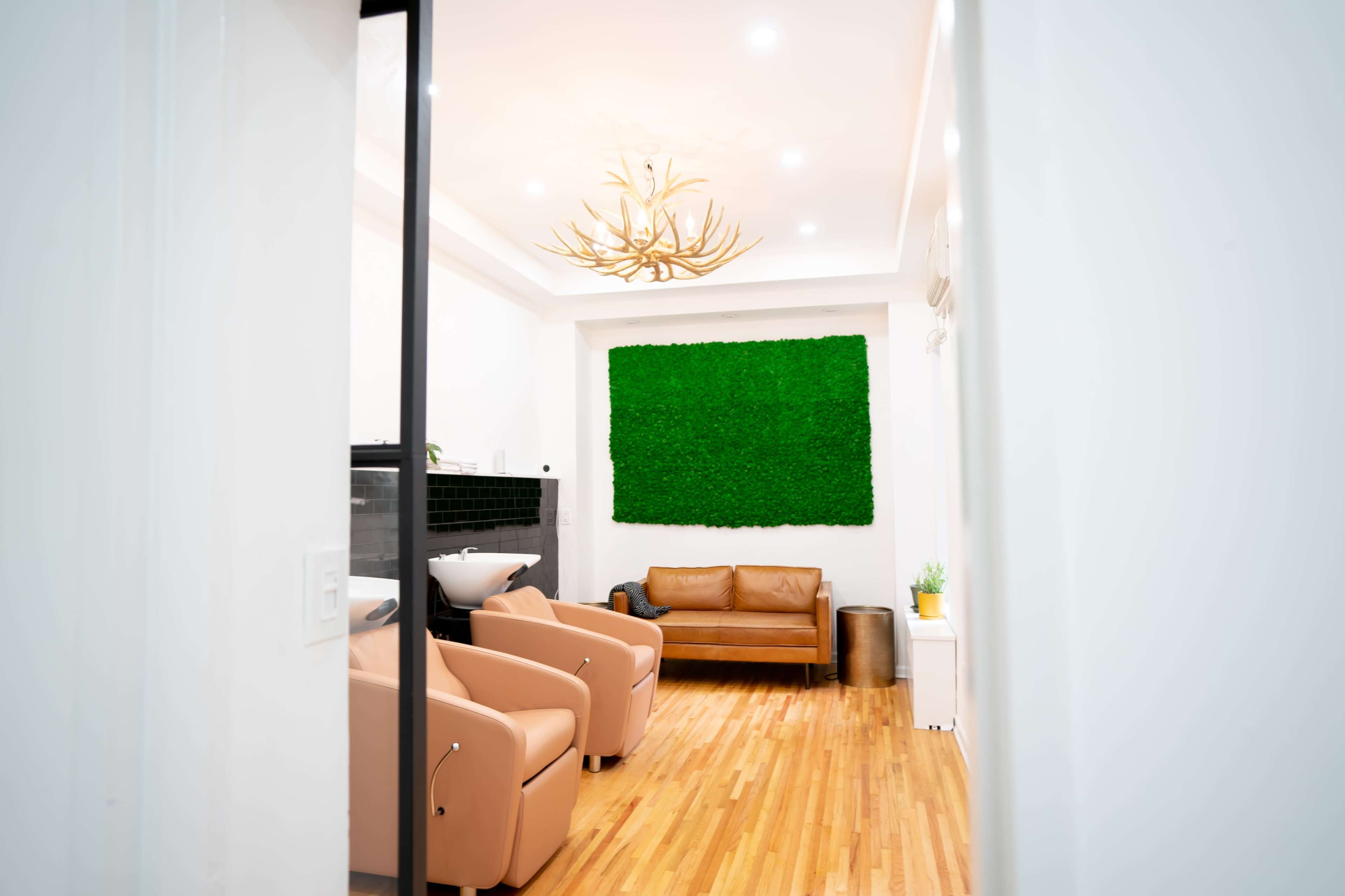 A modern room features a green wall, a brown sofa, and two light-colored chairs, with wooden flooring and a chandelier.