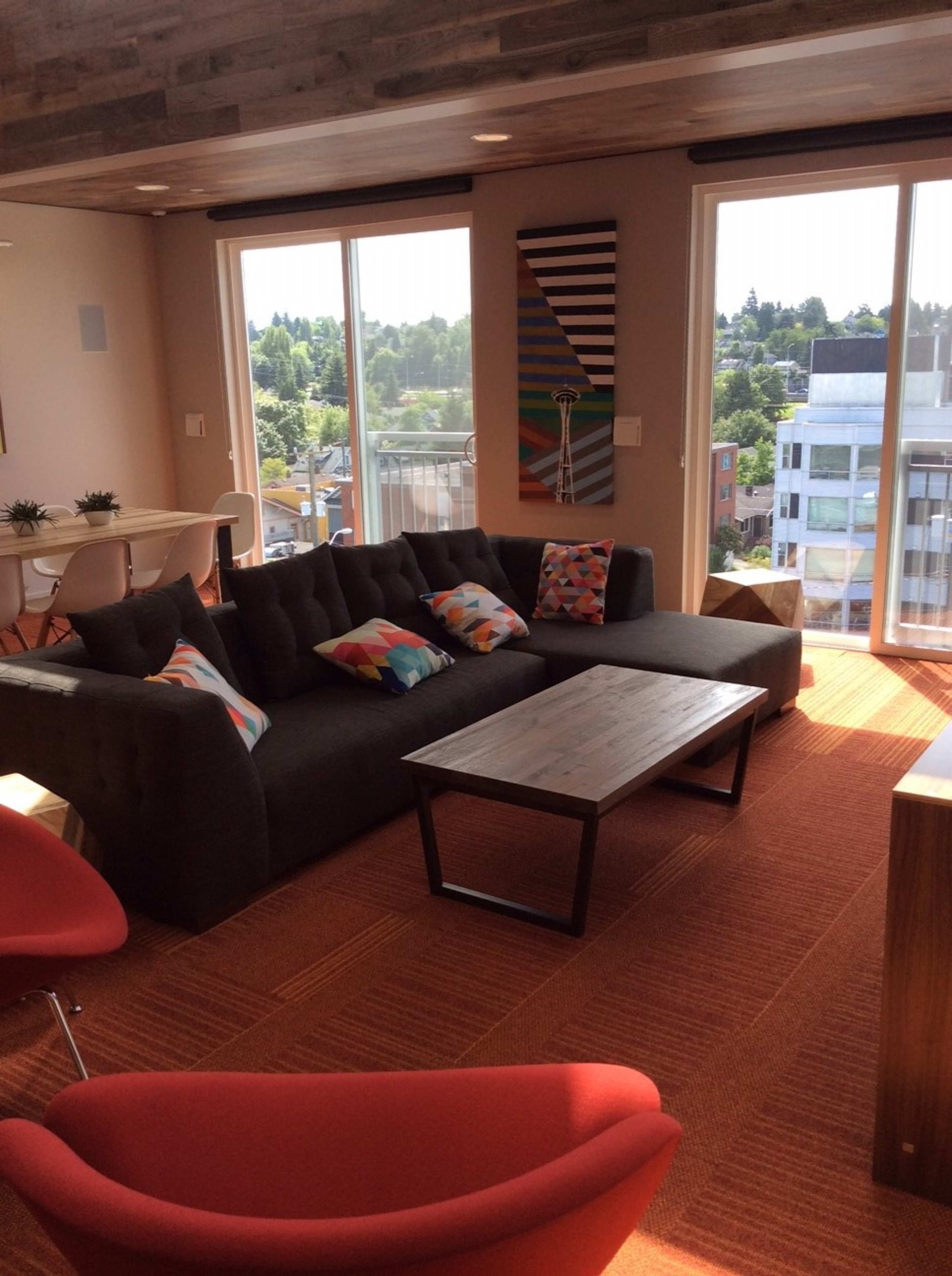 The image shows a modern living area featuring a dark gray couch, a wooden coffee table, and a red chair, with large windows providing a view of a green landscape.