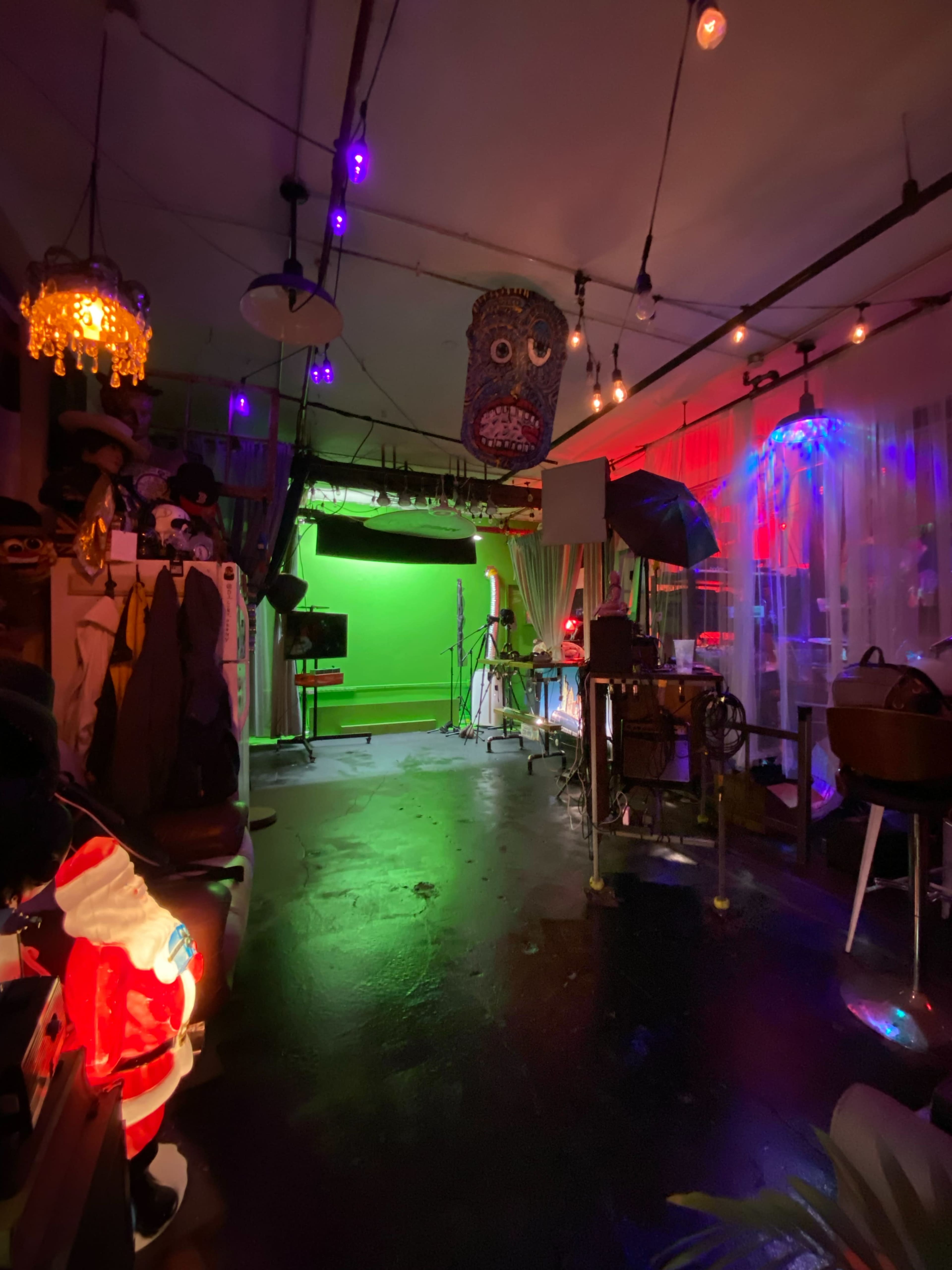 A dimly lit room with a green screen and various props, including a suspended mask-like figure and colorful lighting.