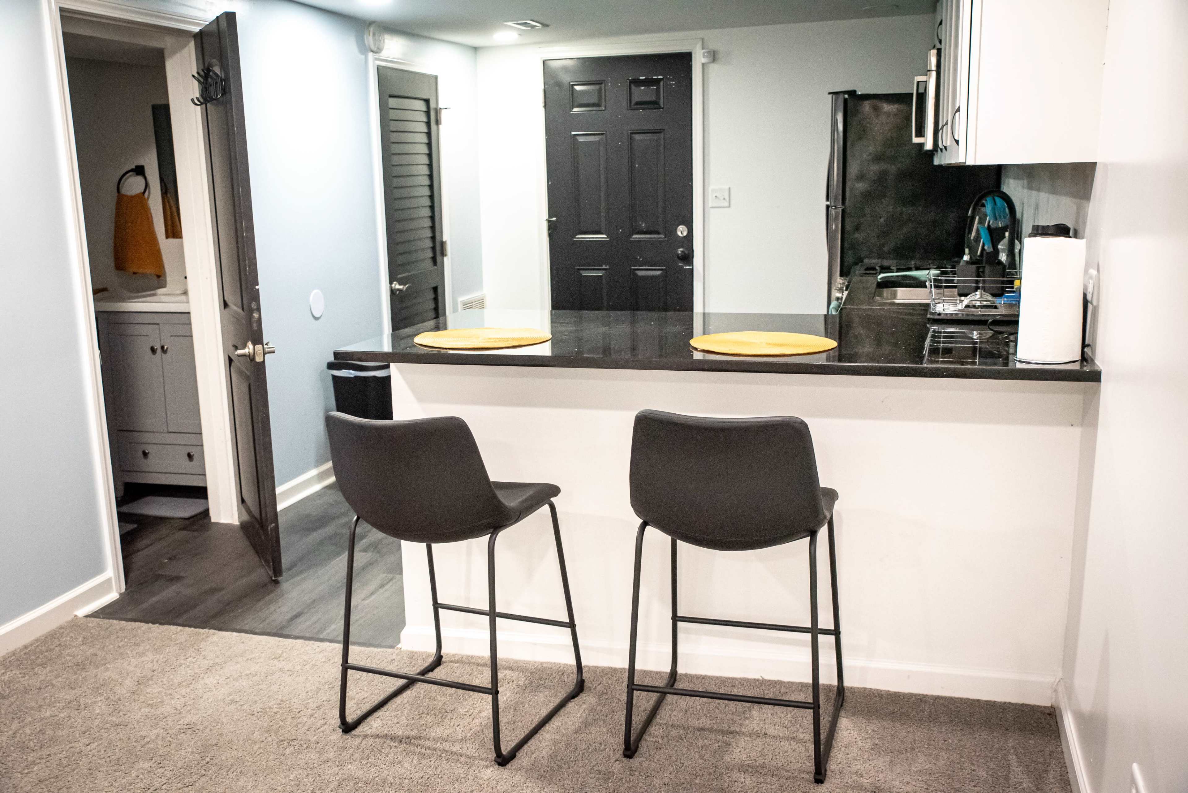 A small kitchen area features a black countertop with two barstools and a doorway leading to a laundry room.