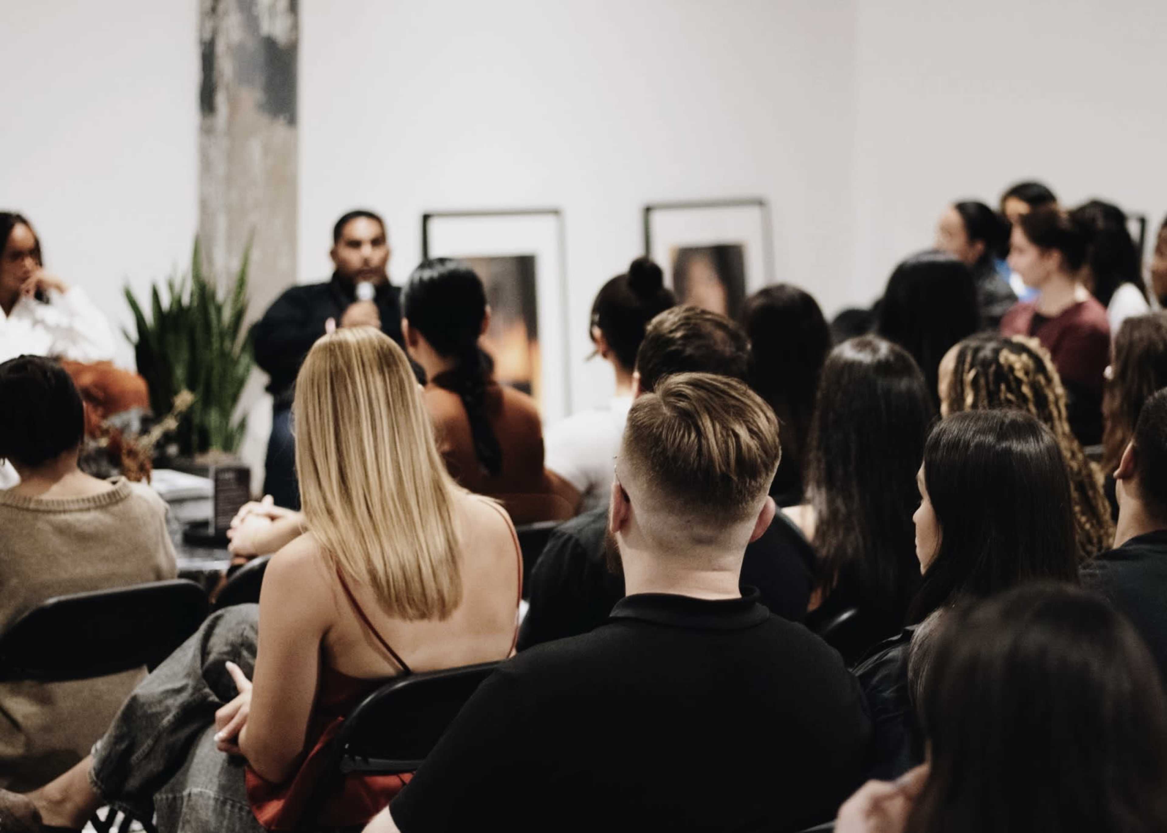 A group of people is seated in a room, facing a speaker who is engaged in conversation, with framed photographs on the walls in the background.